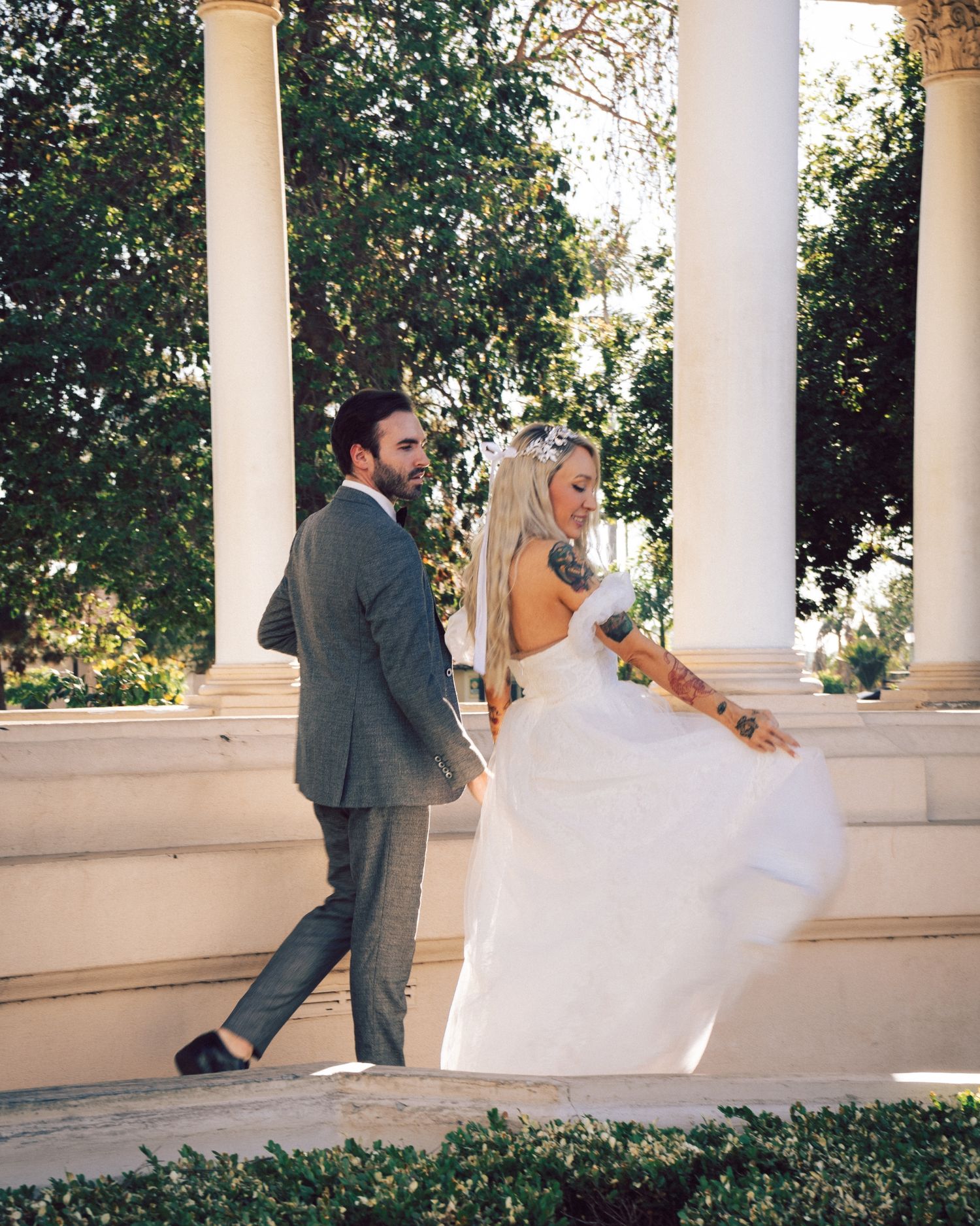 Bride in backless white gown and groom in gray suit walking among white columns at elegant venue.