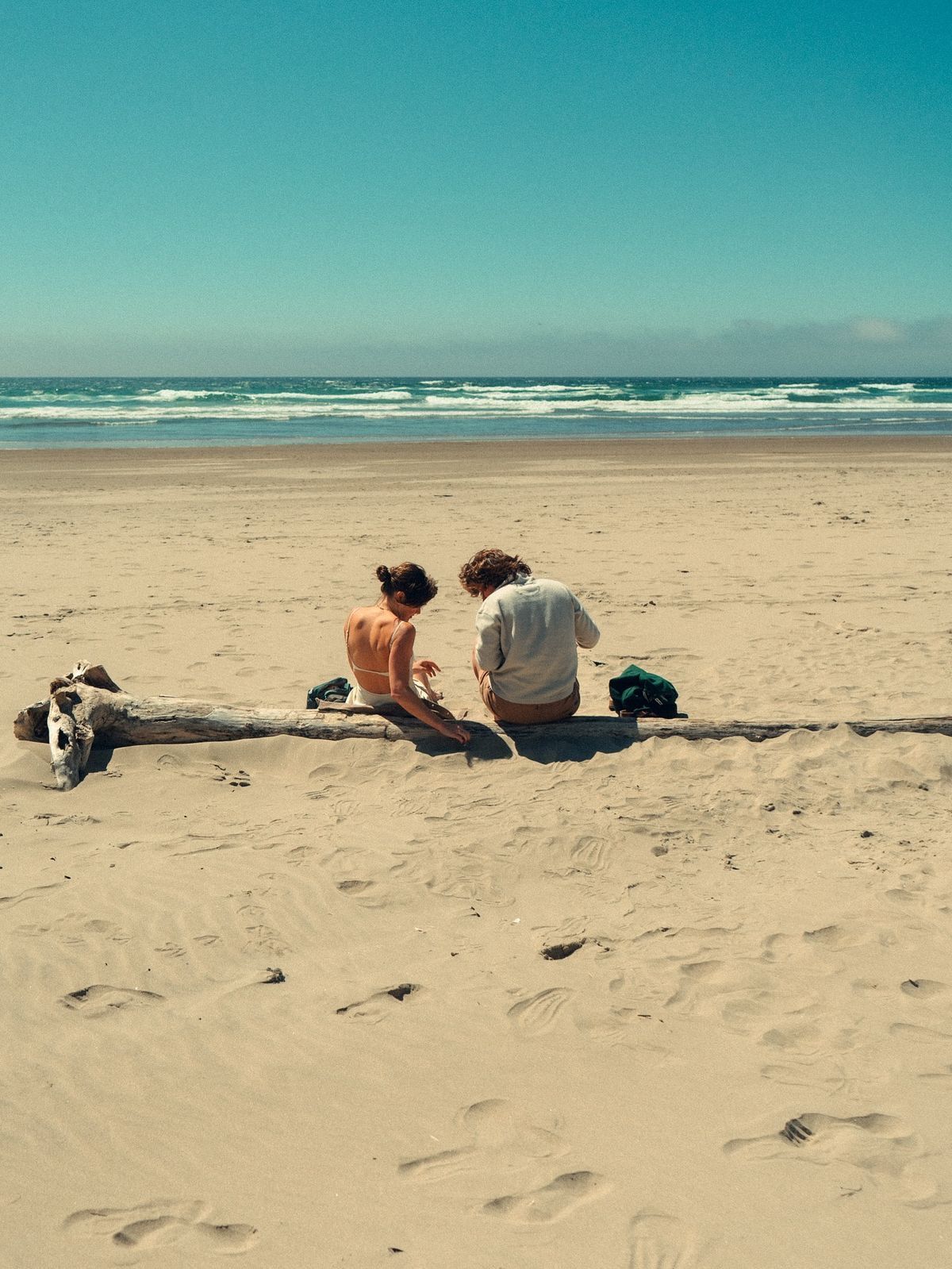 Couple sitting together on sandy beach leaning against driftwood log with ocean waves behind.