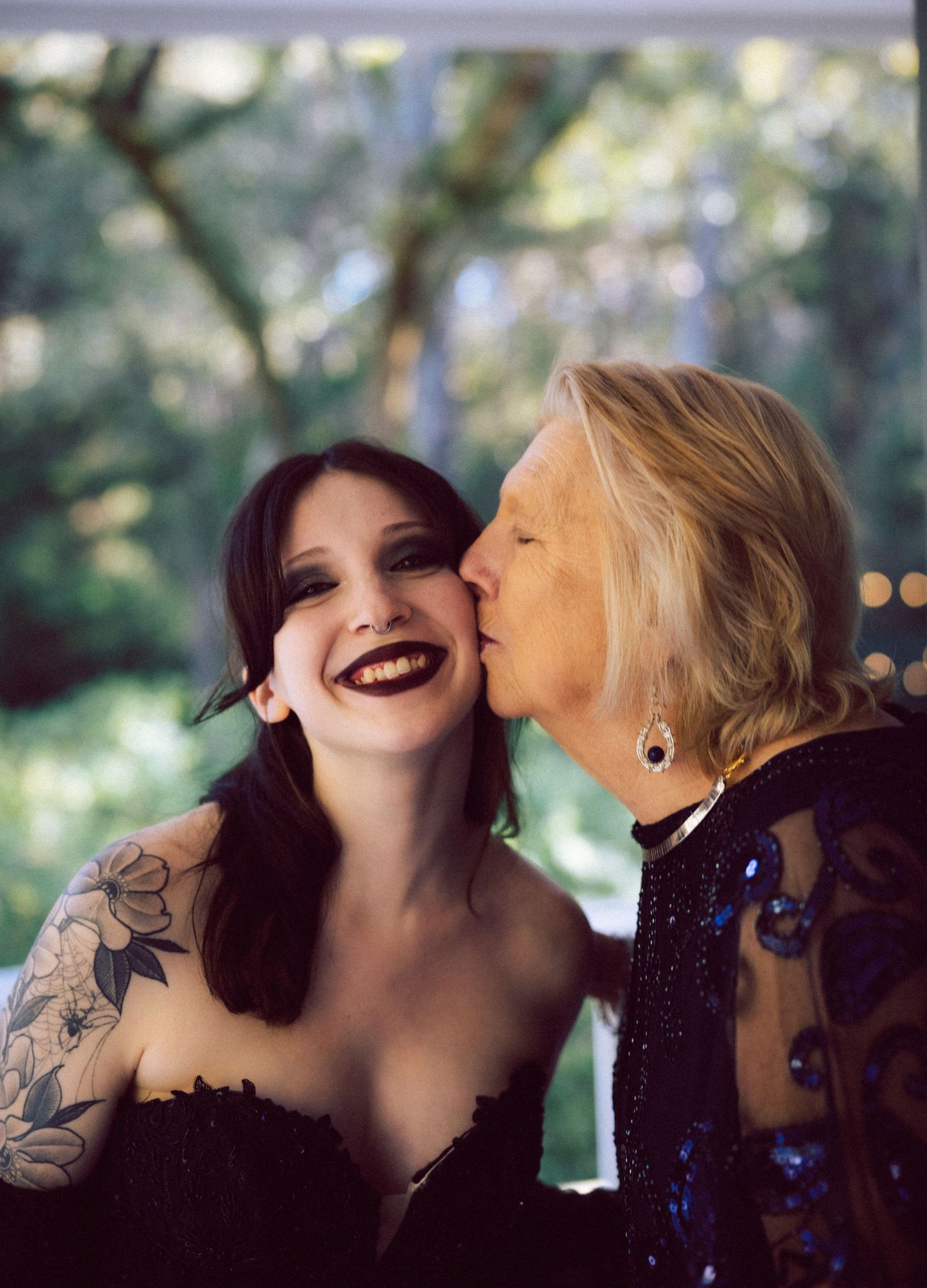 A tattooed bride in a black dress smiles as an elderly woman kisses her cheek outdoors.