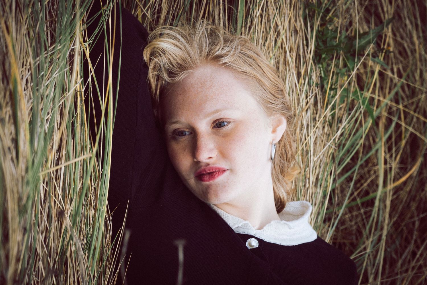 Blonde woman leaning against tall dry grass, wearing a dark cardigan with white collar.