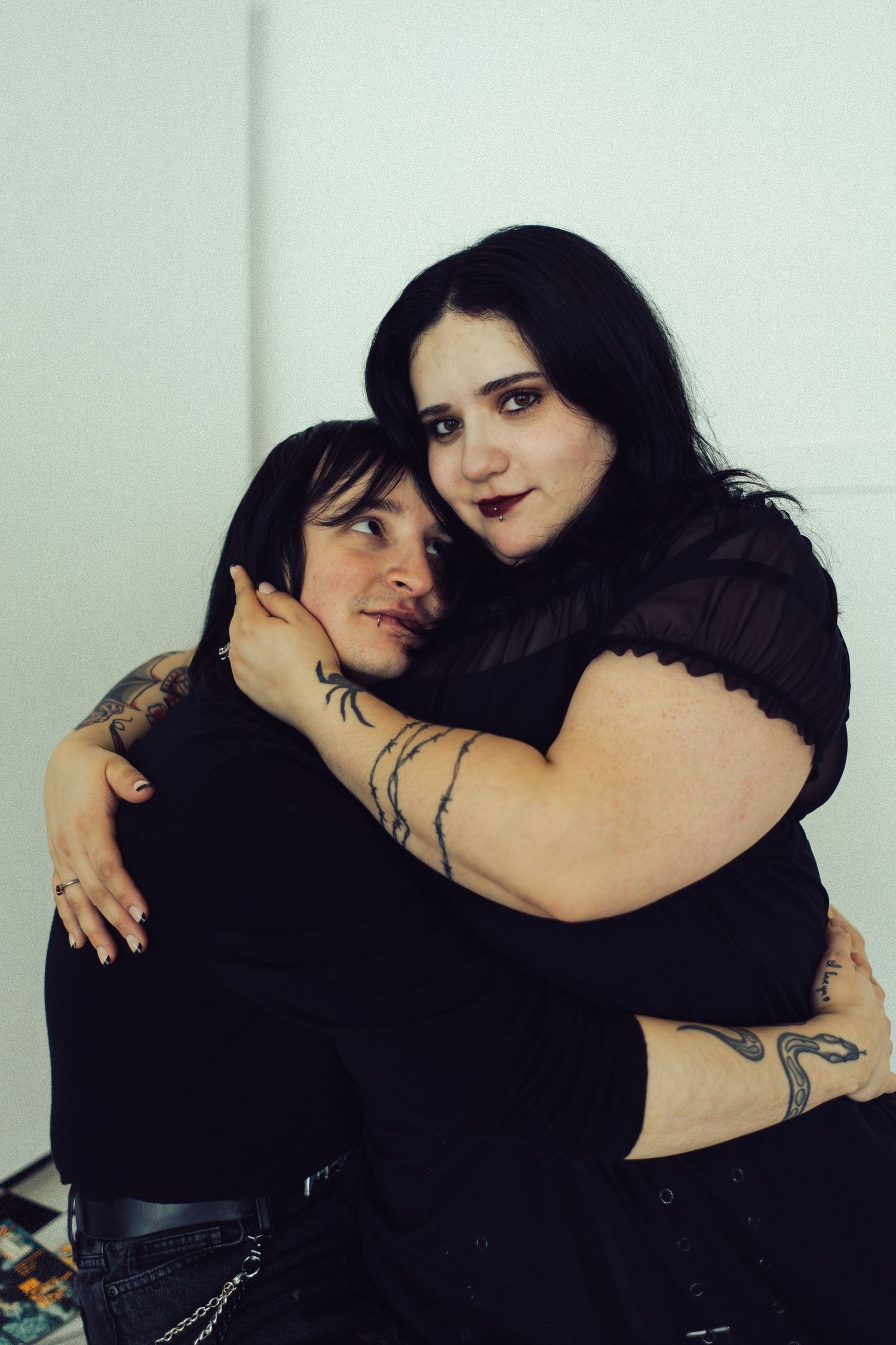 Two women with tattoos sharing a warm embrace against a white wall indoors.