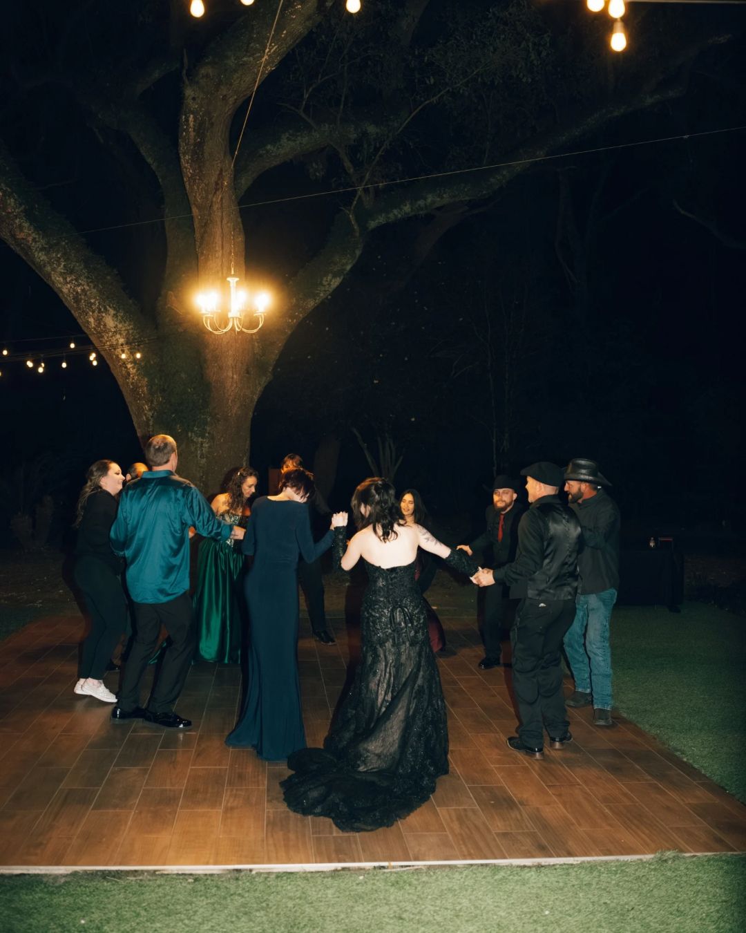 Guests dancing outdoors at night under string lights and chandelier beside a large oak tree.