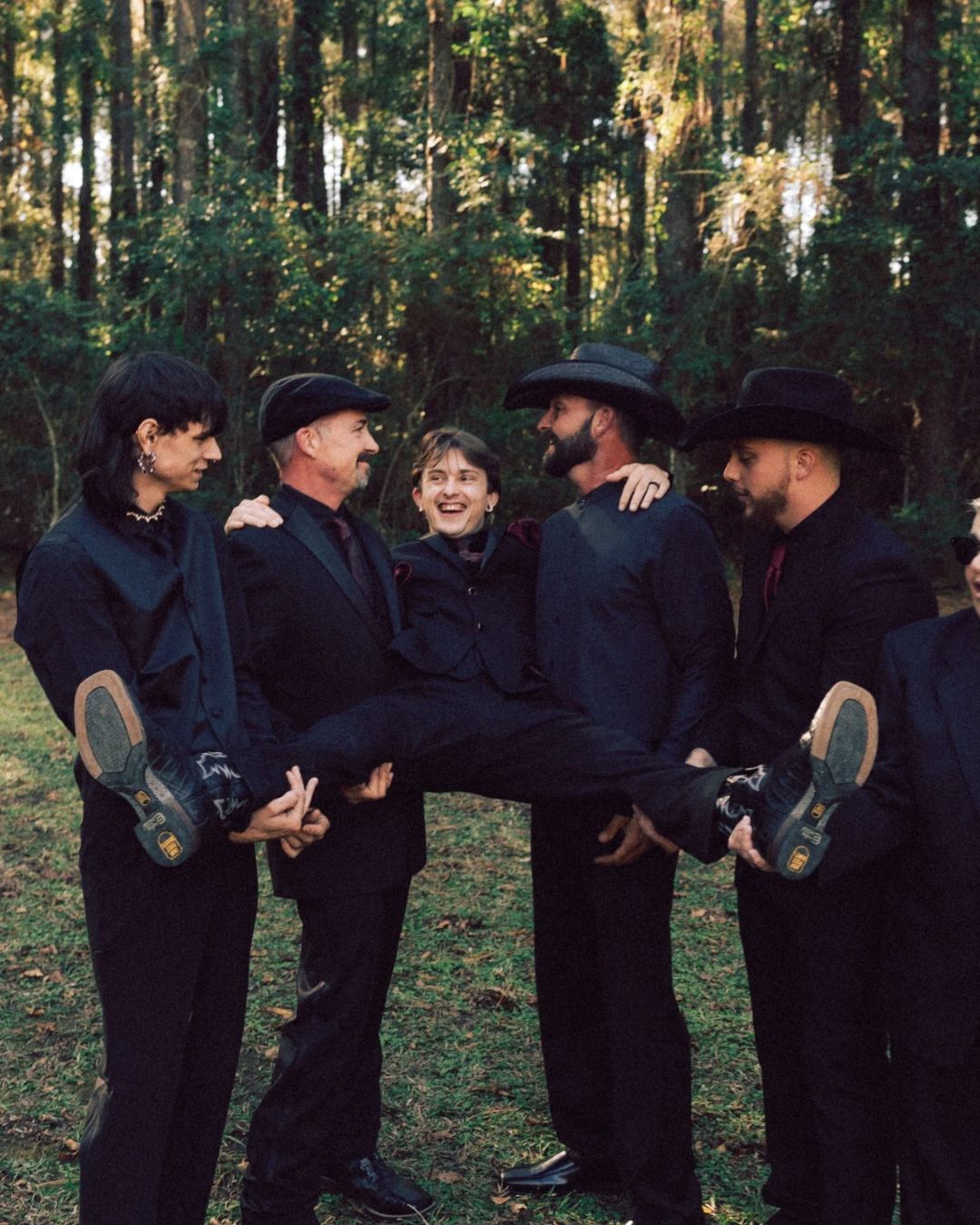 Group of men in all-black outfits and cowboy hats laughing and lifting one person in a forest.