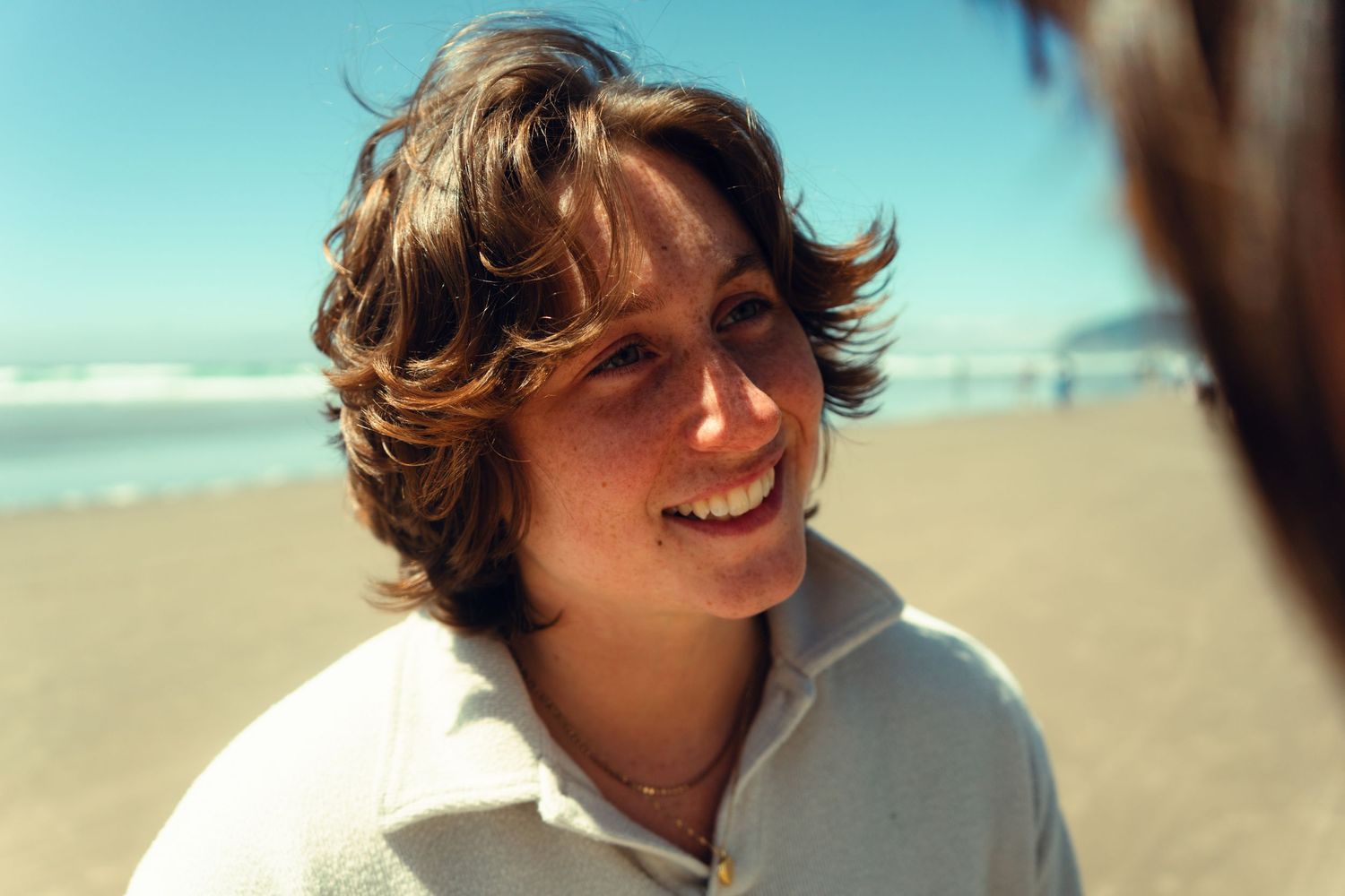 A smiling person with tousled hair enjoys a sunny day at the beach in a white shirt.