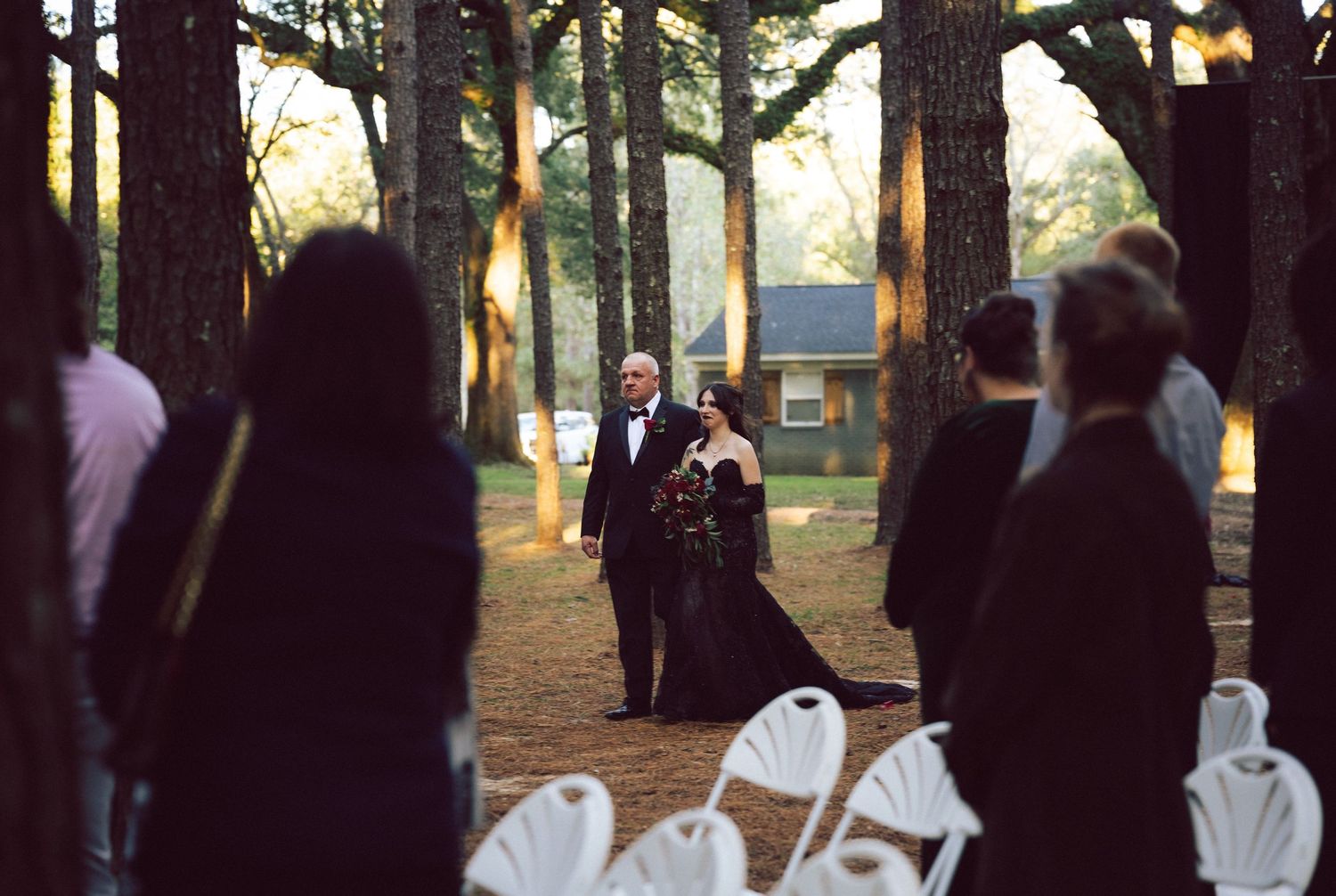 Bride in black gown walks down forest aisle with escort at golden hour pine tree wedding ceremony.