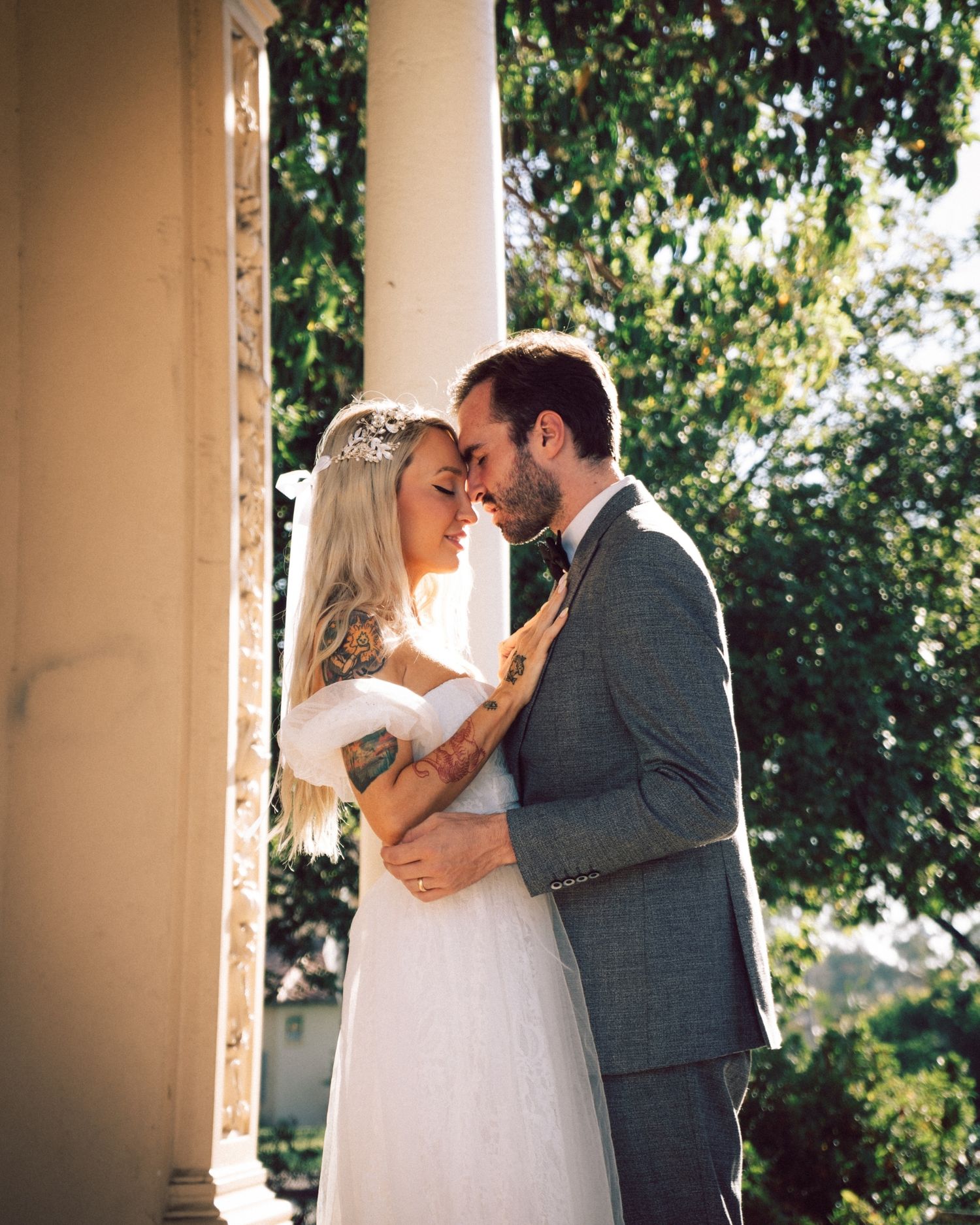 A bride and groom share a tender moment between white columns surrounded by lush green trees.