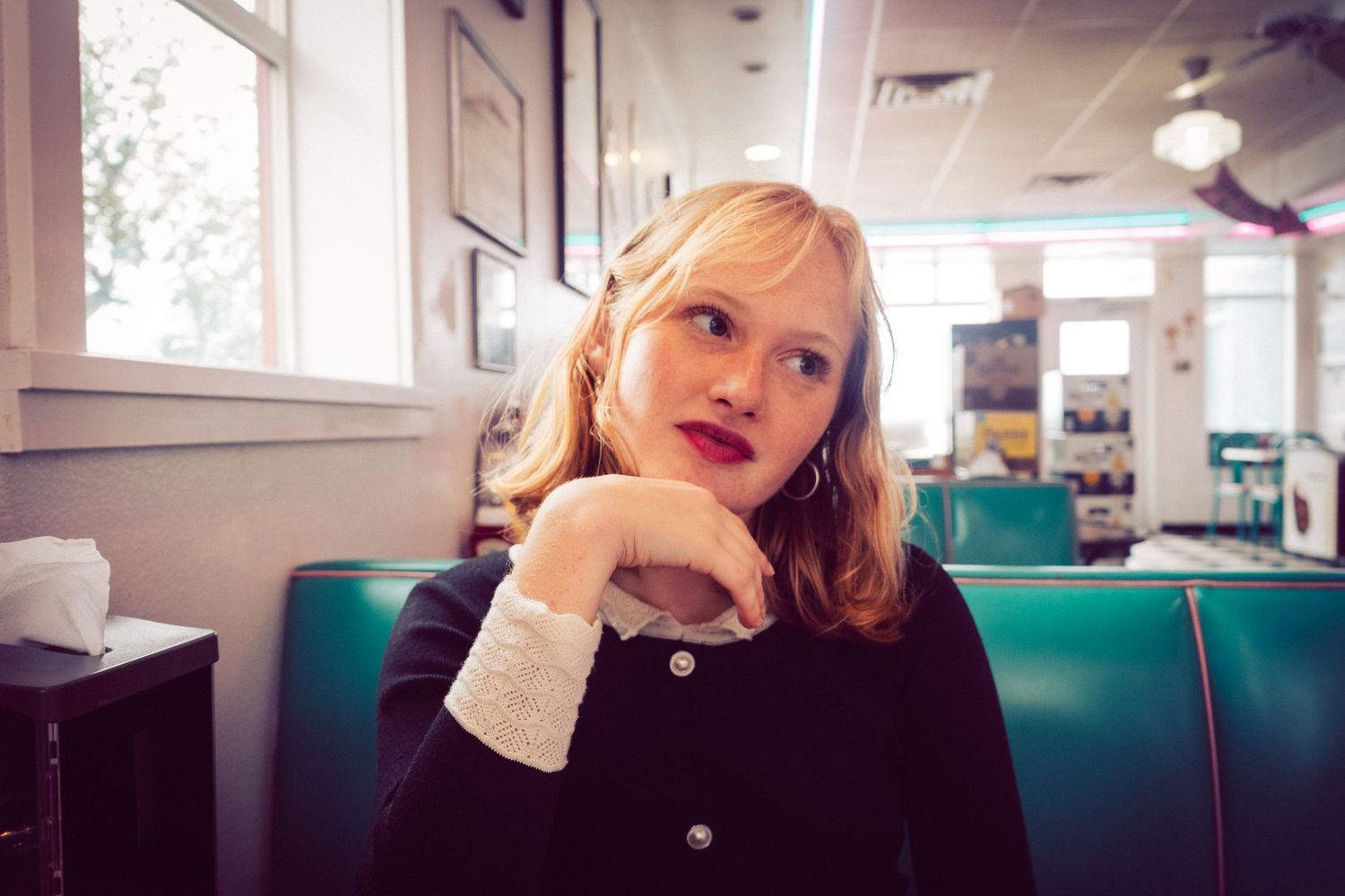 Young blonde woman with red lips sitting in a vintage diner booth looking away.