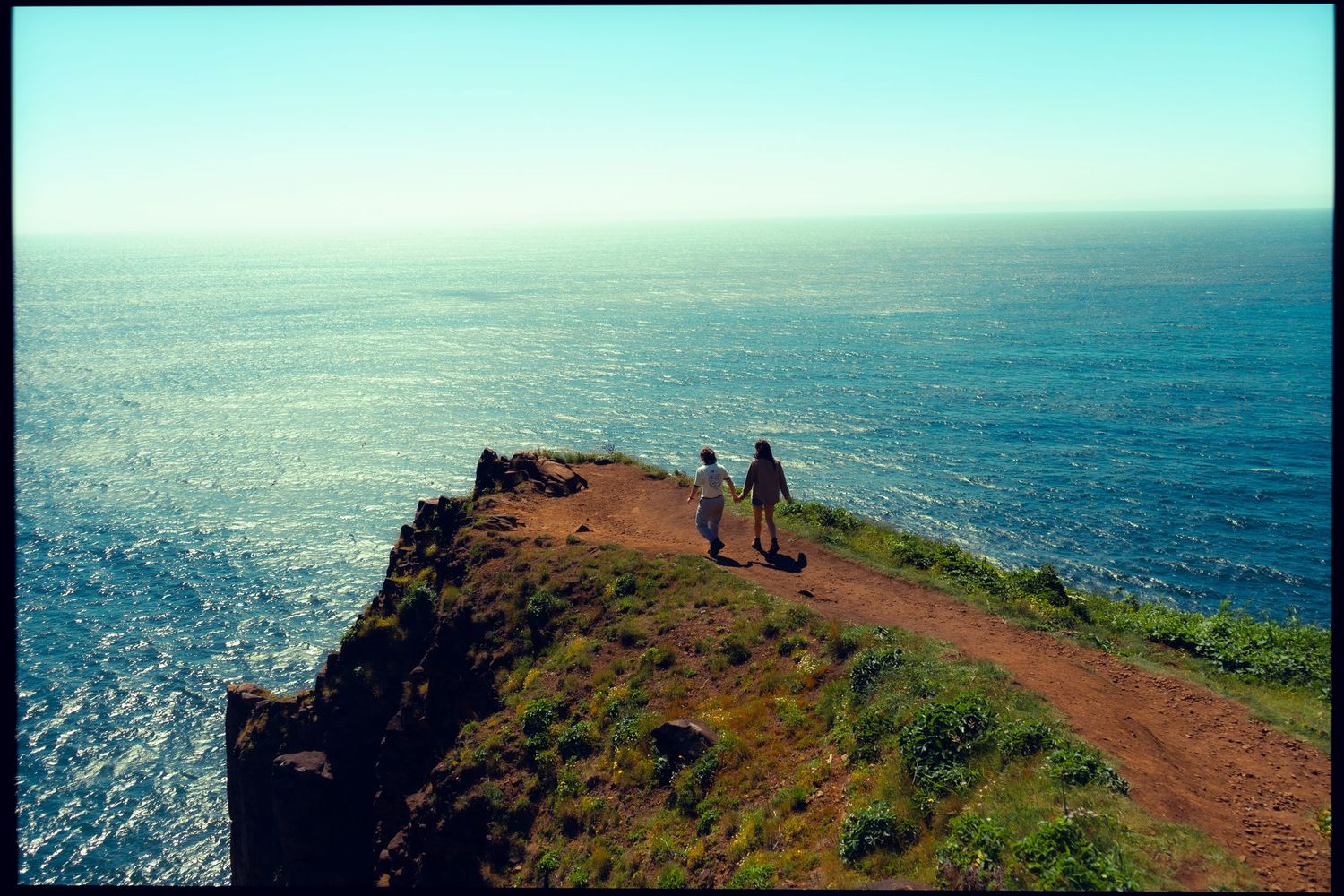 Couple holding hands on a dramatic coastal cliff overlooking a vast blue ocean on a clear day.