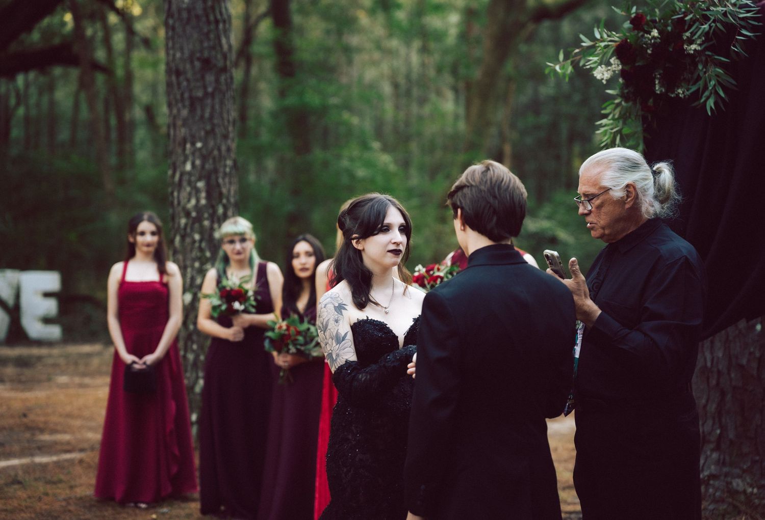 Gothic wedding ceremony in pine forest with bride, groom, officiant, and bridesmaids in burgundy gowns.