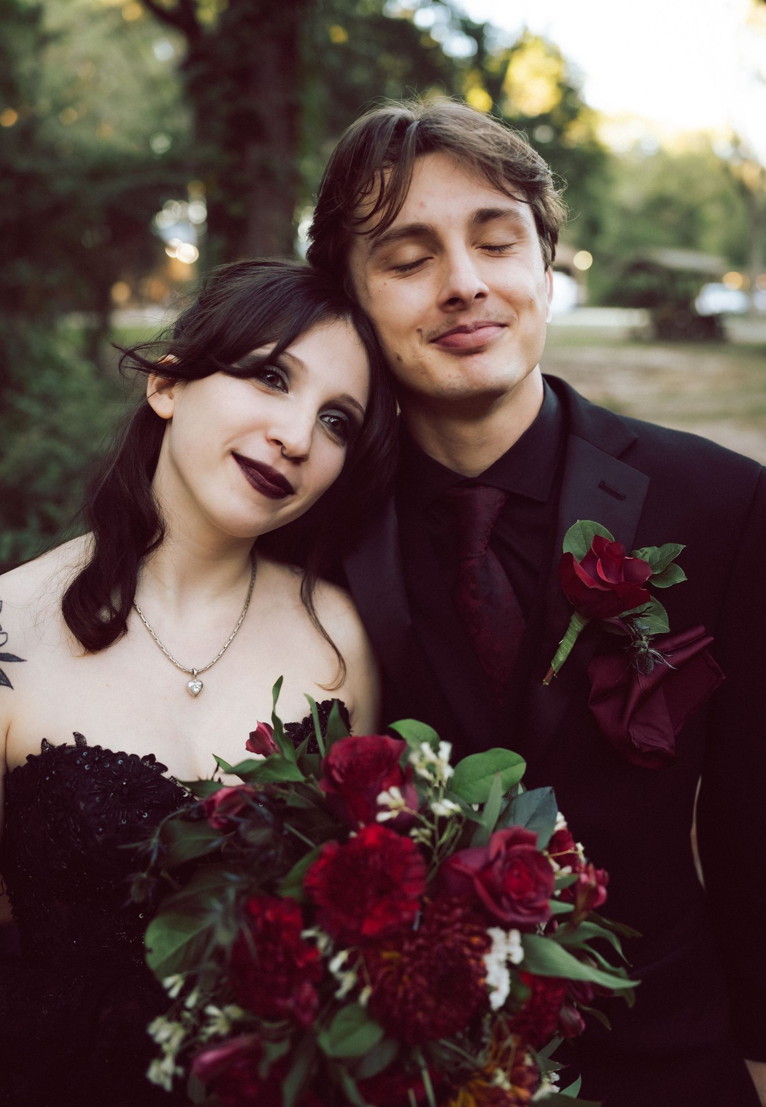 Gothic wedding couple posing with dark red bouquet, bride in black dress with dark lipstick, groom in black suit.