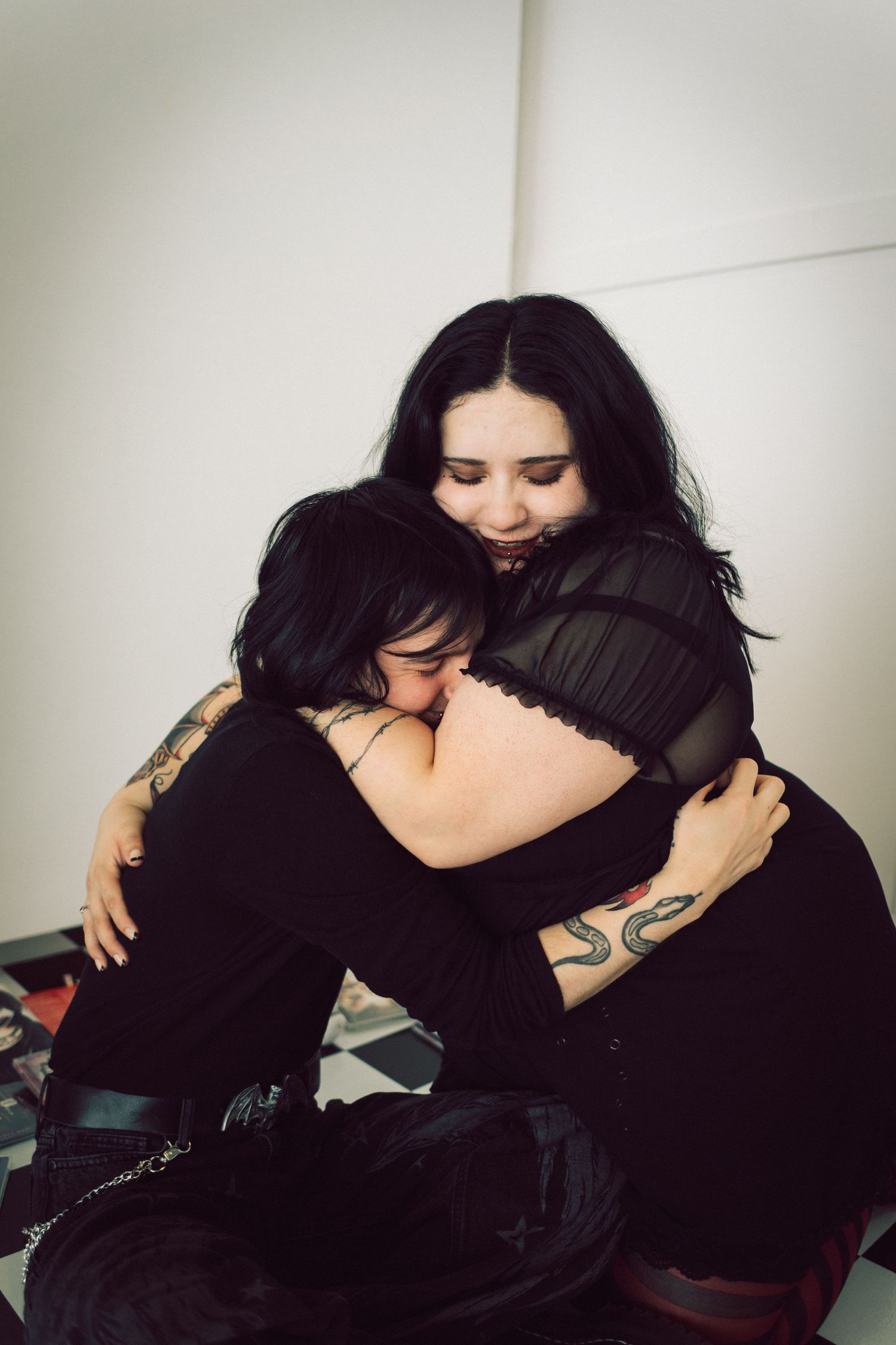 Two women with dark hair and tattoos sharing a warm embrace while wearing black clothing.