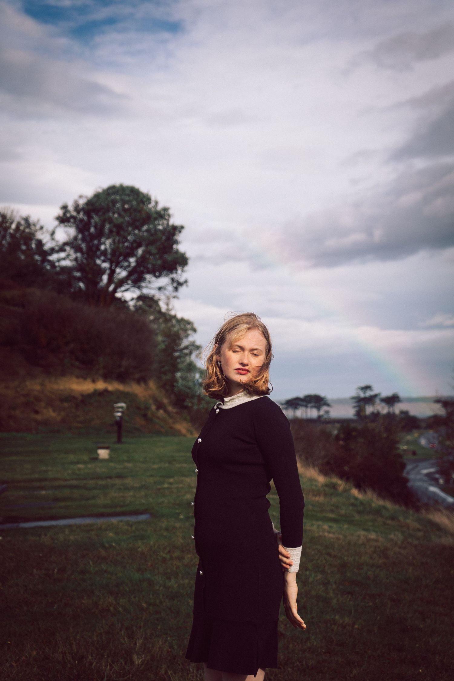 Young woman in black dress standing on a grassy hillside with a rainbow and dramatic cloudy sky.