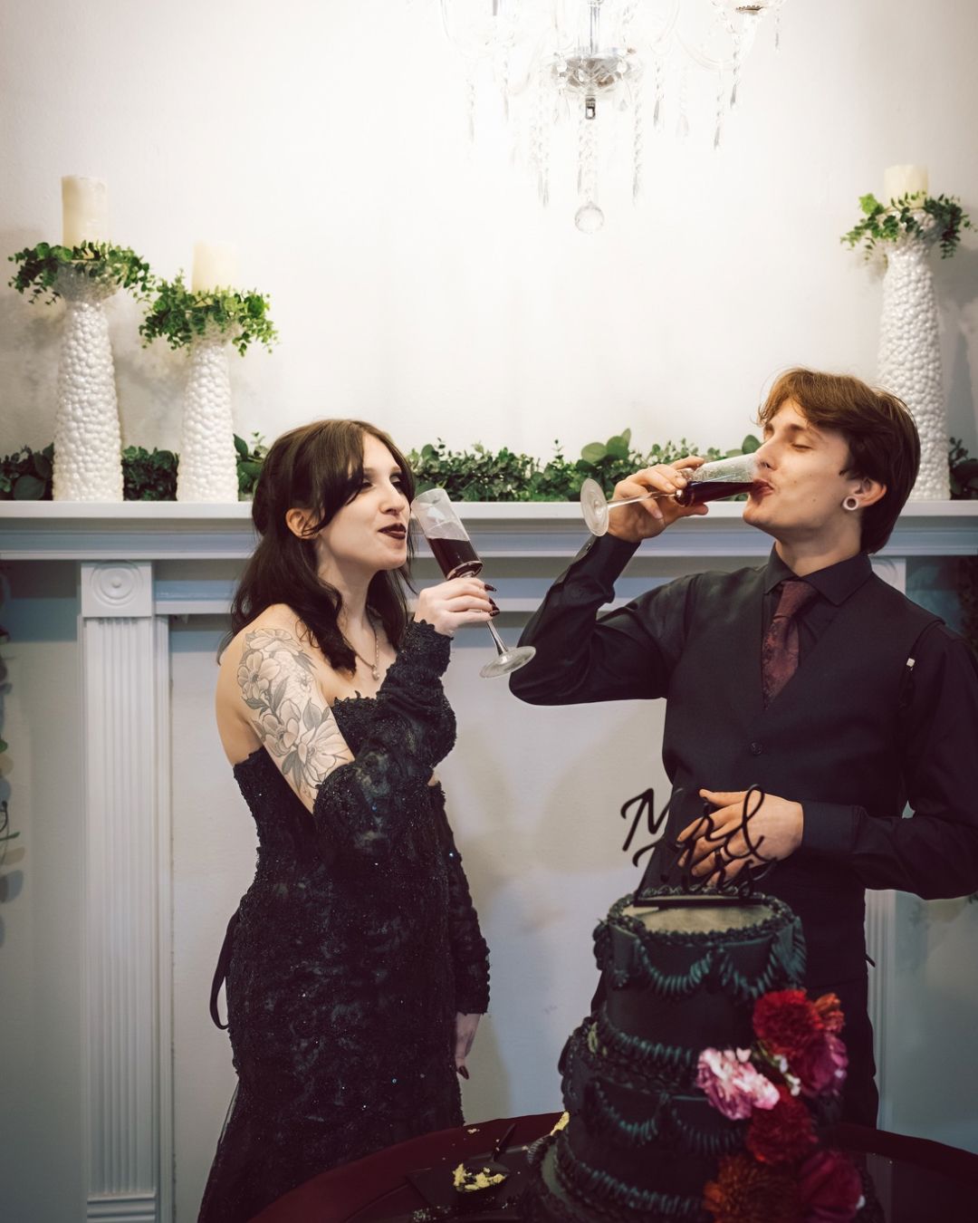 Couple in black wedding attire drinking red wine beside a dark gothic tiered wedding cake.