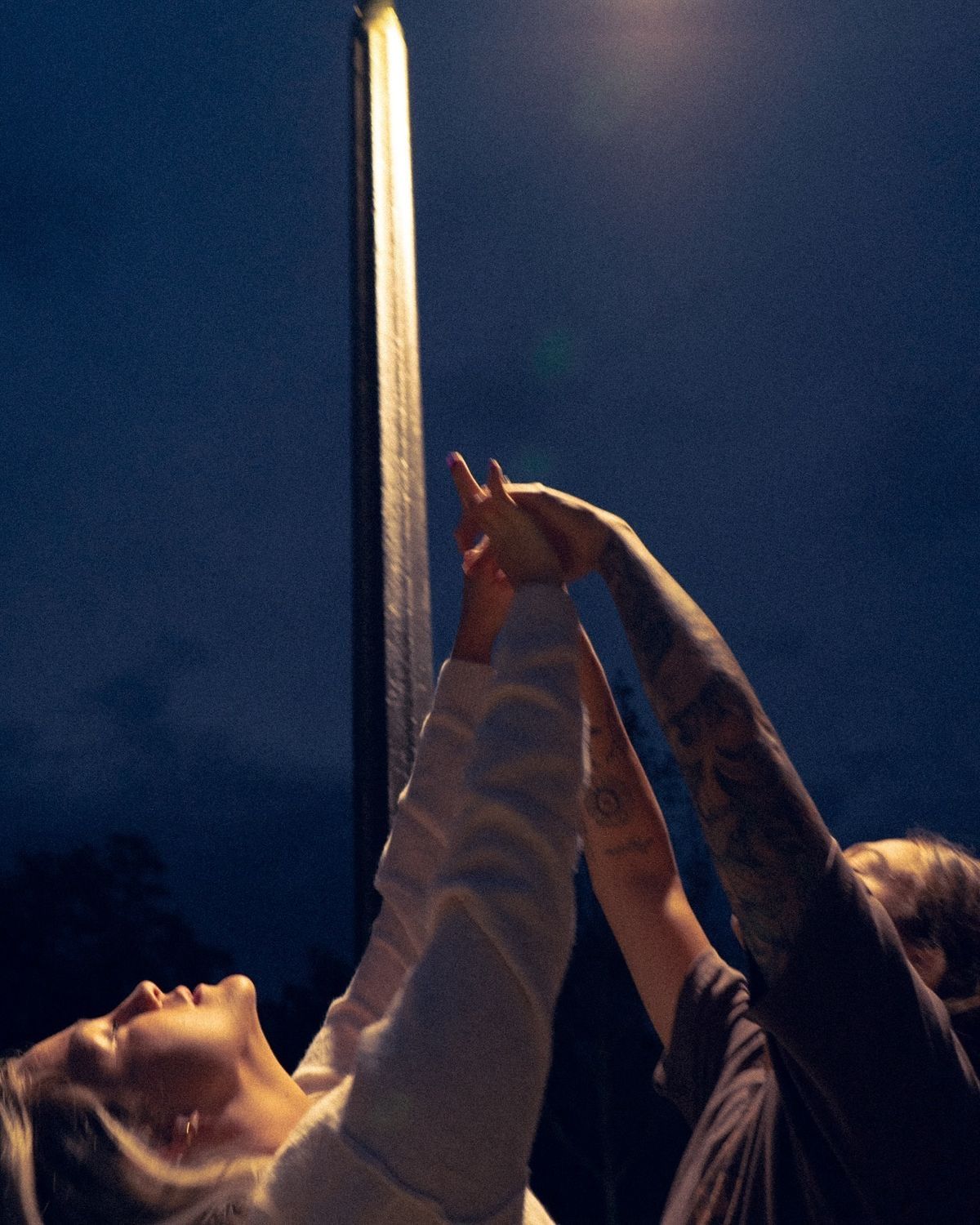 Two people holding hands and pointing up at a street light against a night sky.