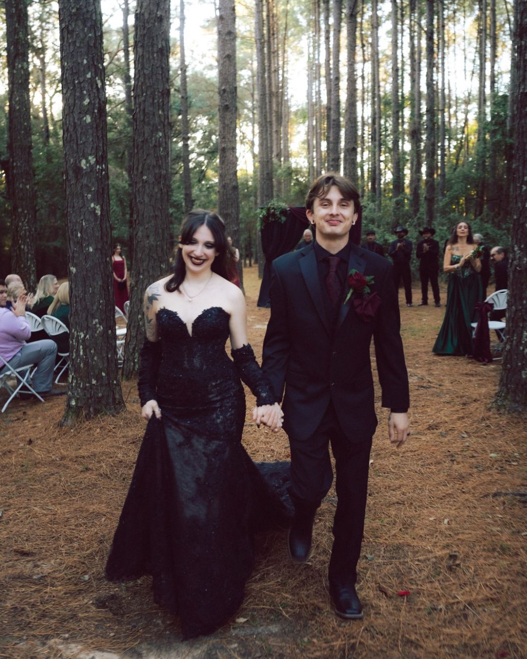 A couple in black formal attire walks hand in hand through a pine forest wedding ceremony.