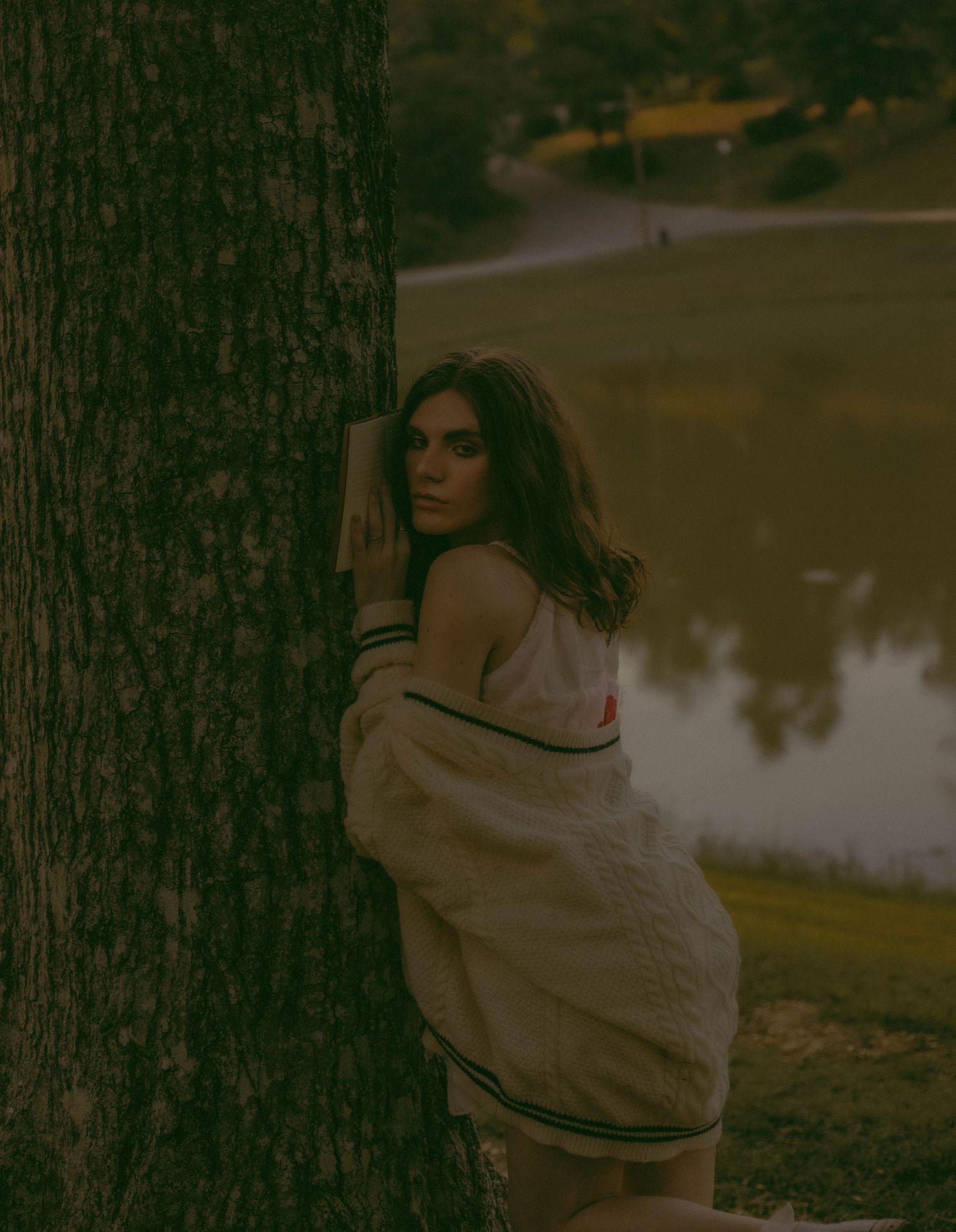 Young woman leaning against a large tree trunk near a lake at dusk in a moody outdoor portrait.