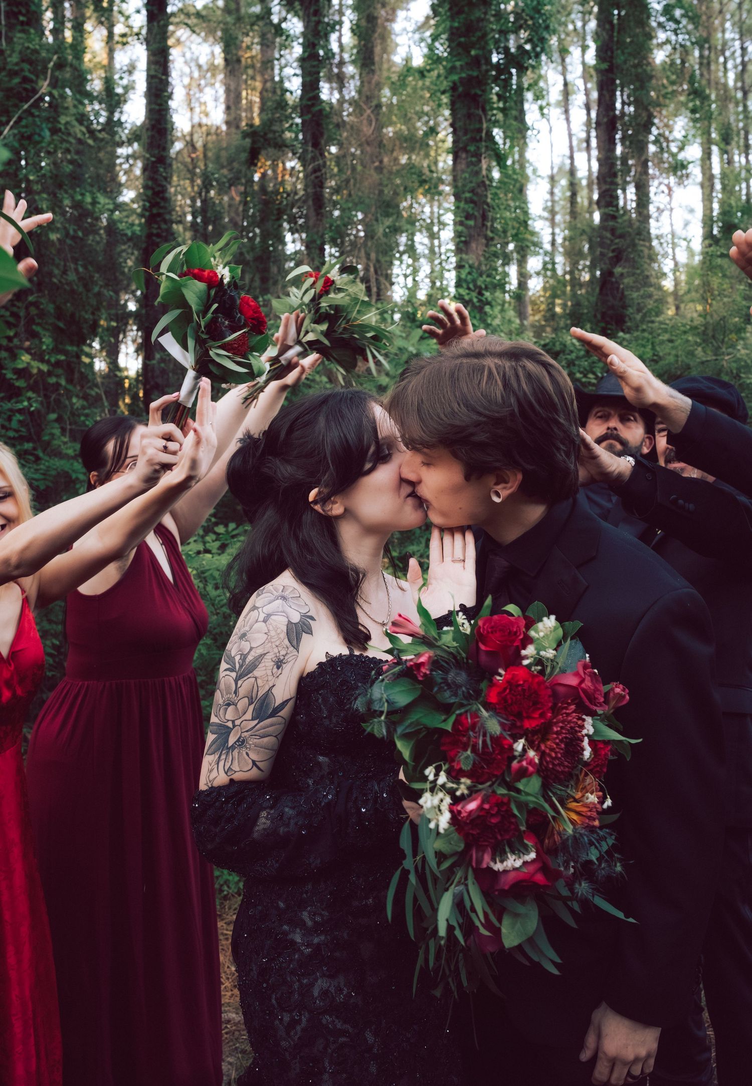 Gothic bride in black lace gown kissing groom holding red bouquet surrounded by wedding party in forest.