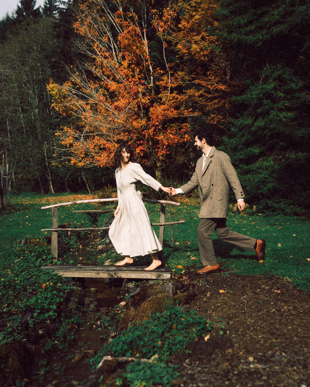 A couple holds hands while crossing a rustic wooden bridge surrounded by vibrant autumn foliage.