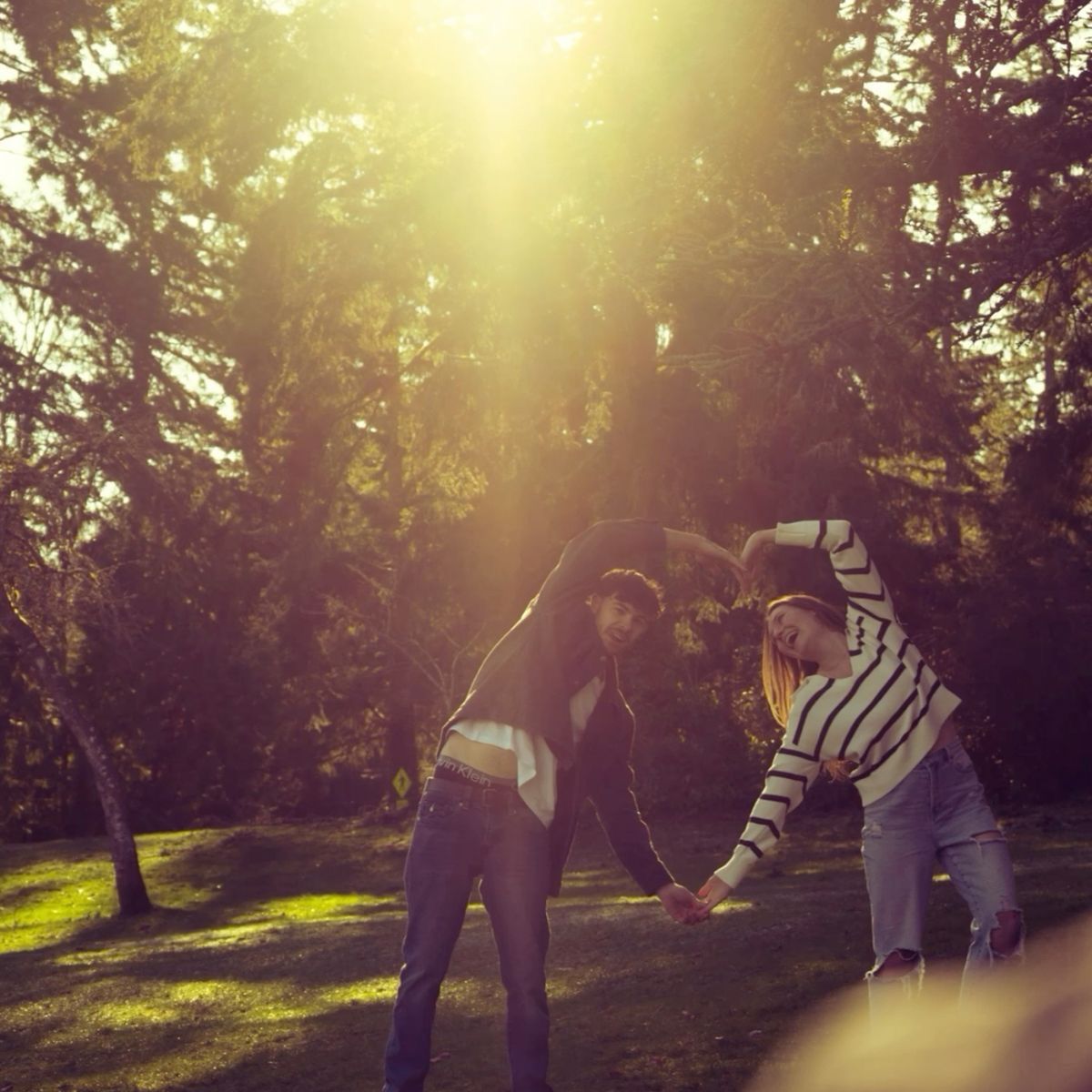Two friends playfully posing in golden sunlight filtering through tall trees in a park.