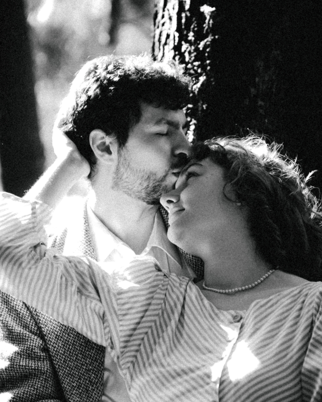 Black and white romantic couple sharing a kiss near a tree in soft sunlight.