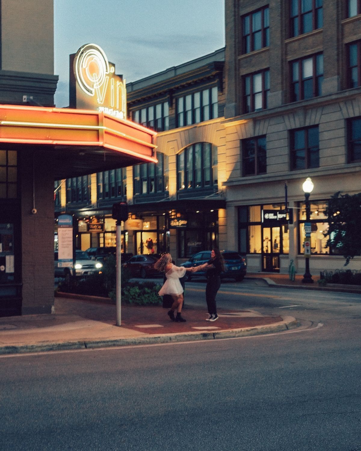 A couple dances joyfully outside a vintage theater with glowing neon signs at dusk.