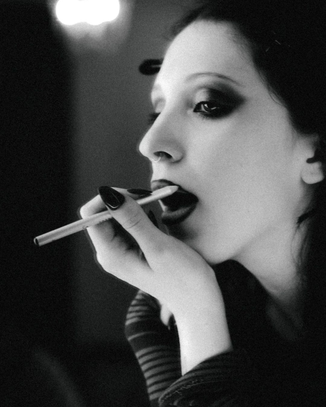 Black and white portrait of gothic woman applying dark lipstick with dramatic smoky eye makeup.