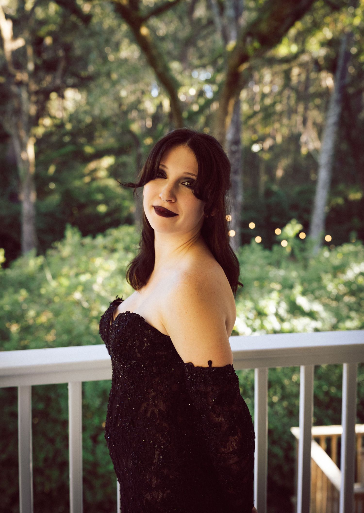 Bride in black off-shoulder lace gown poses on wooden balcony surrounded by lush green trees.