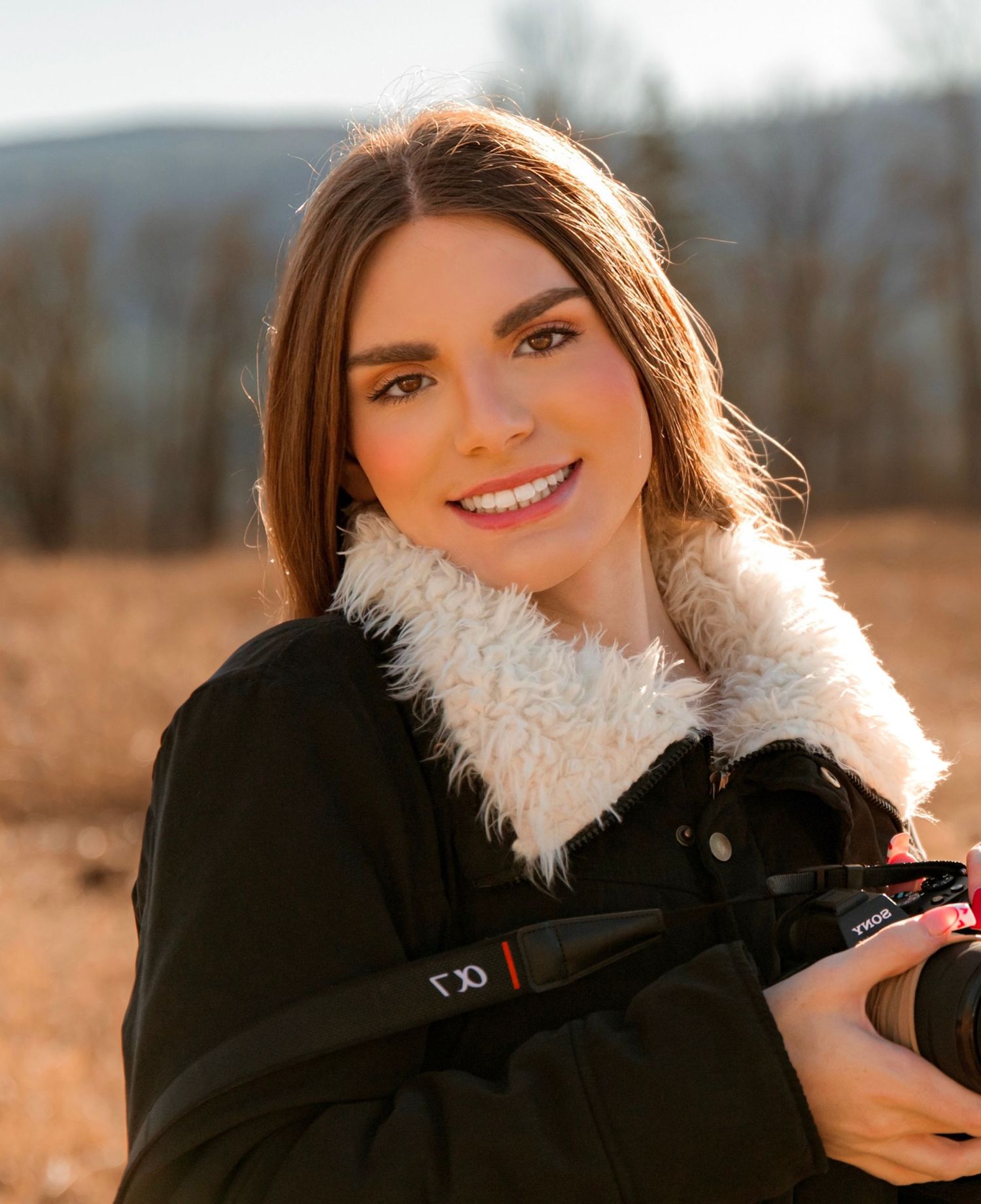 Smiling young woman holding a camera outdoors wearing a black coat with white fur collar in golden sunlight.