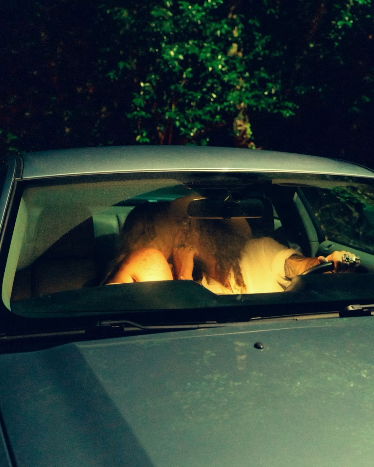 Two people embracing inside a car viewed through windshield at night with dark foliage background.