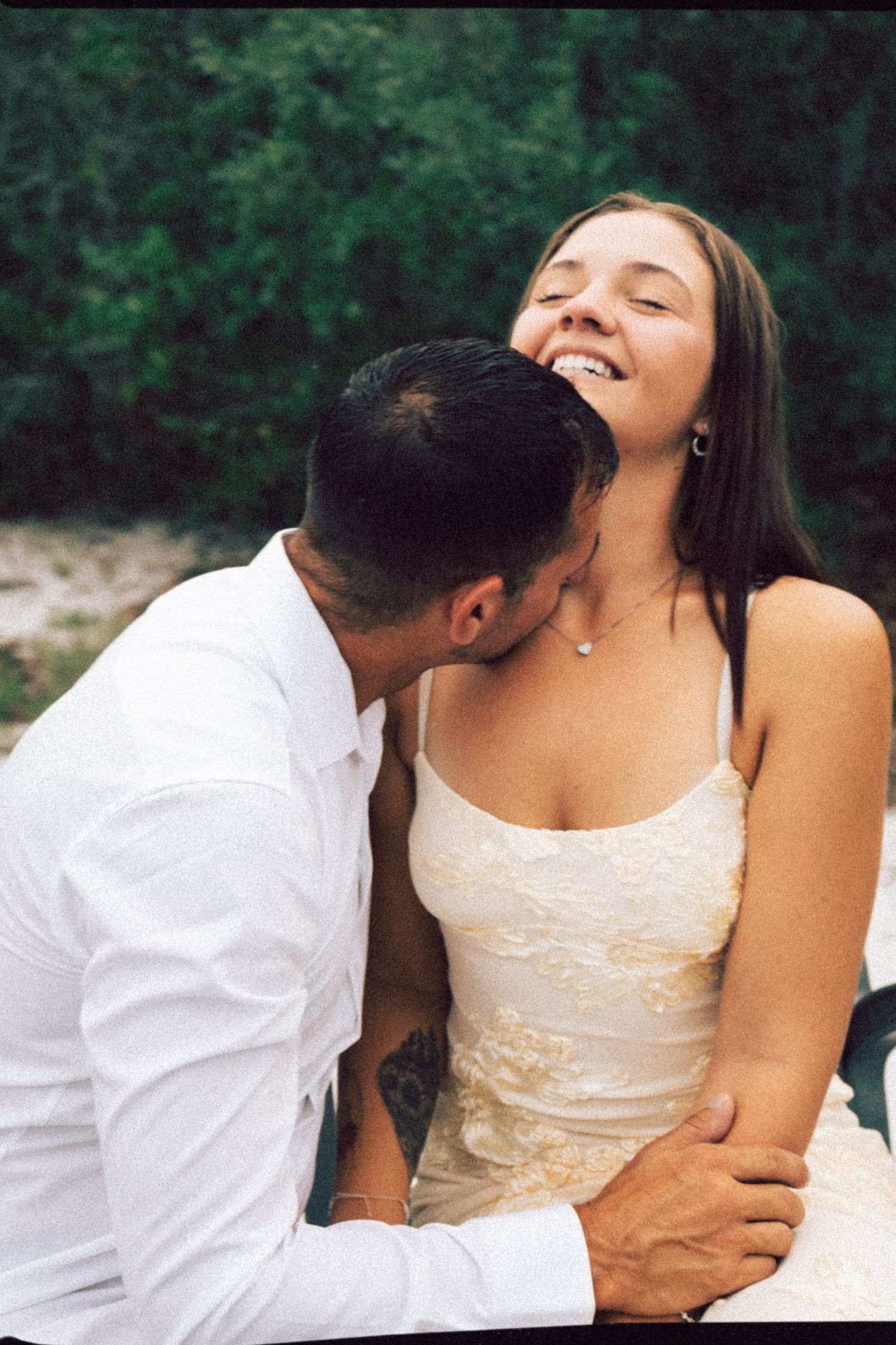 Man kissing womans neck while she laughs joyfully outdoors near green foliage.