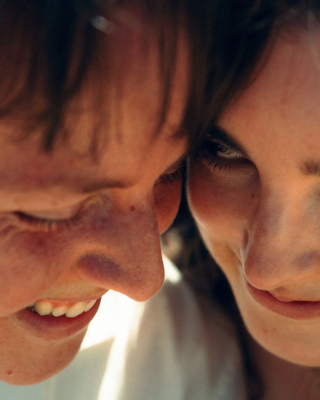 Close-up candid portrait of two people smiling and leaning heads together in warm natural light.