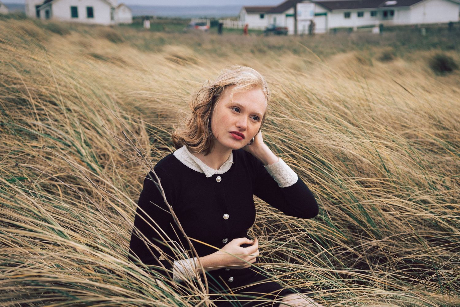 Young blonde woman sitting among tall golden grass in a coastal field, wearing a dark navy dress.