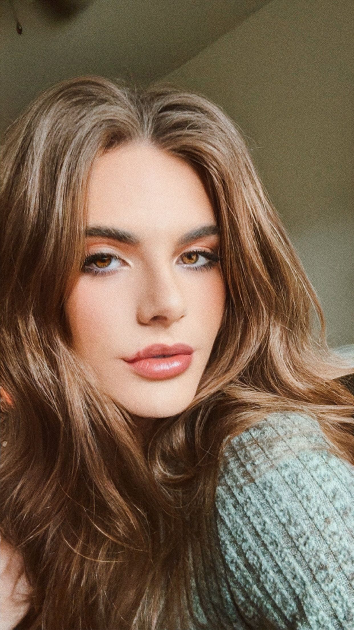 Young woman with long brown hair and natural makeup wearing a teal sweater in a close-up selfie.