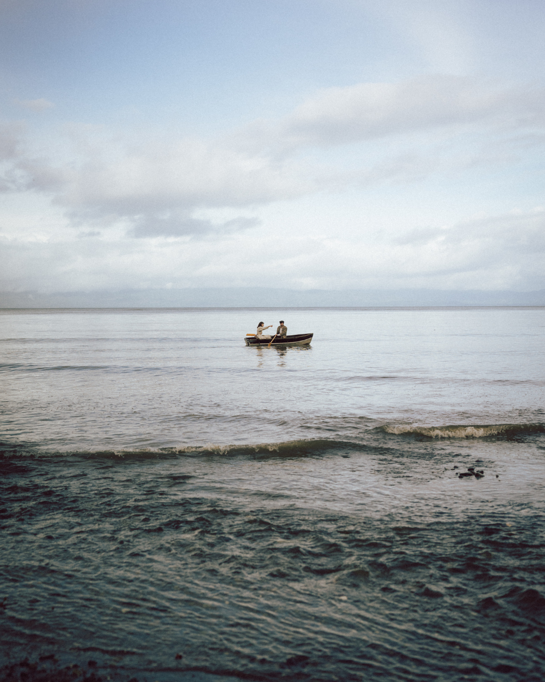 Two people row a small boat on a calm, expansive ocean under a cloudy sky.