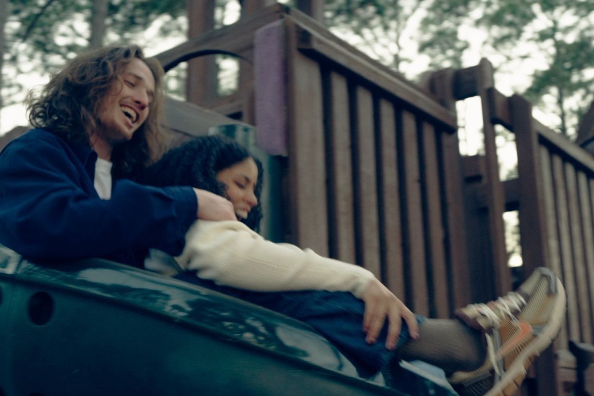 Two people laughing and cuddling on a playground slide surrounded by trees.