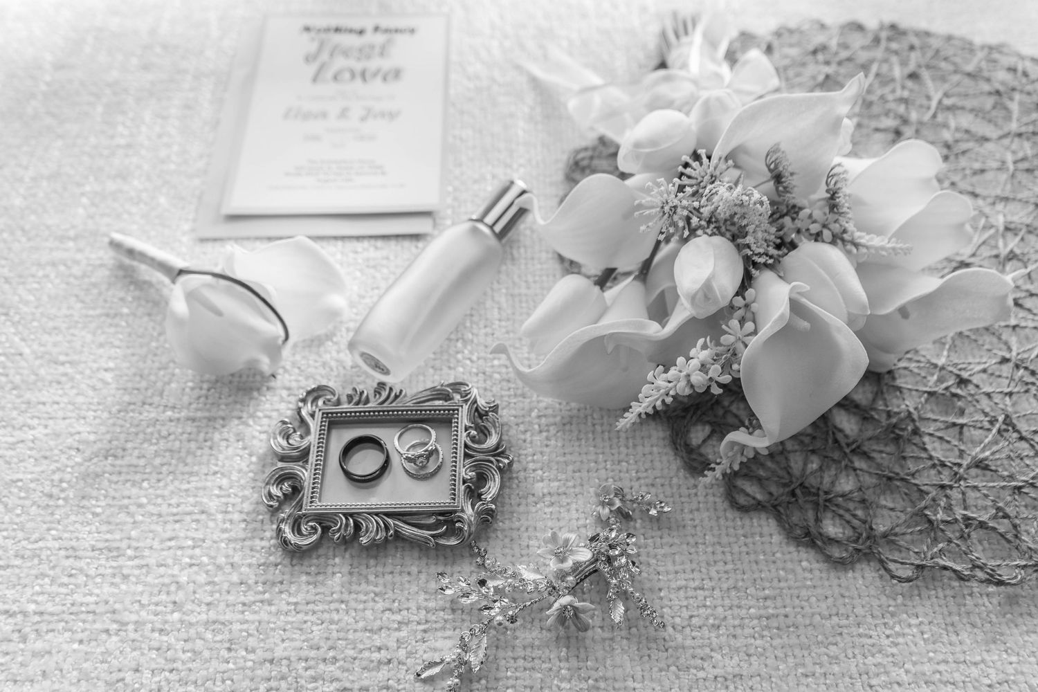 Black and white flat lay of wedding rings, bridal bouquet, perfume, and invitation on textured fabric.