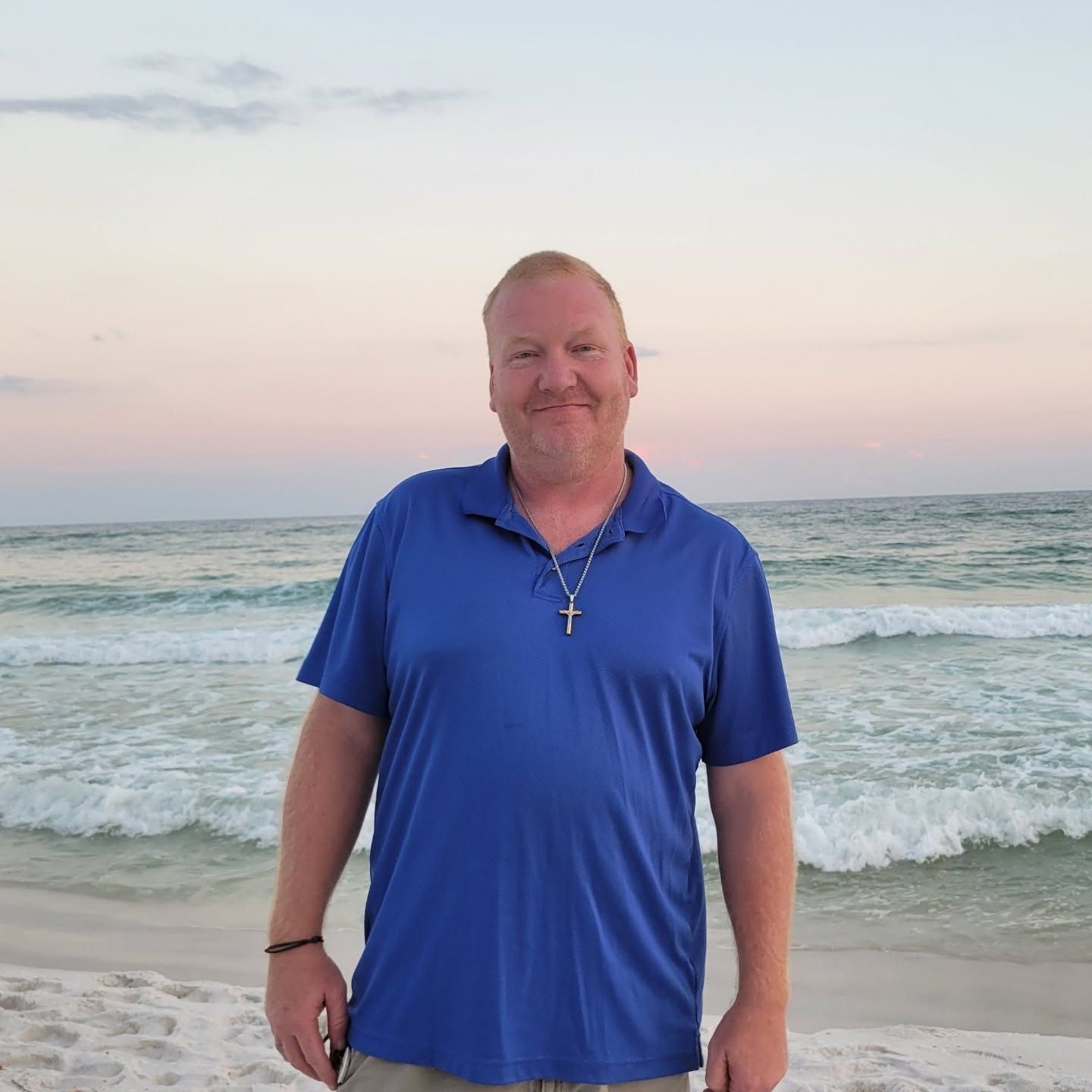 Smiling man in blue polo shirt standing on a sandy beach at sunset with ocean waves behind him.