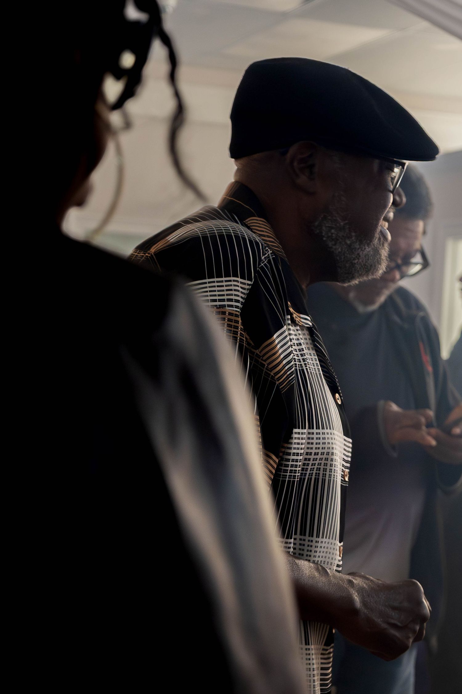 A bearded man wearing a black beret and patterned shirt stands in a dimly lit indoor gathering space.