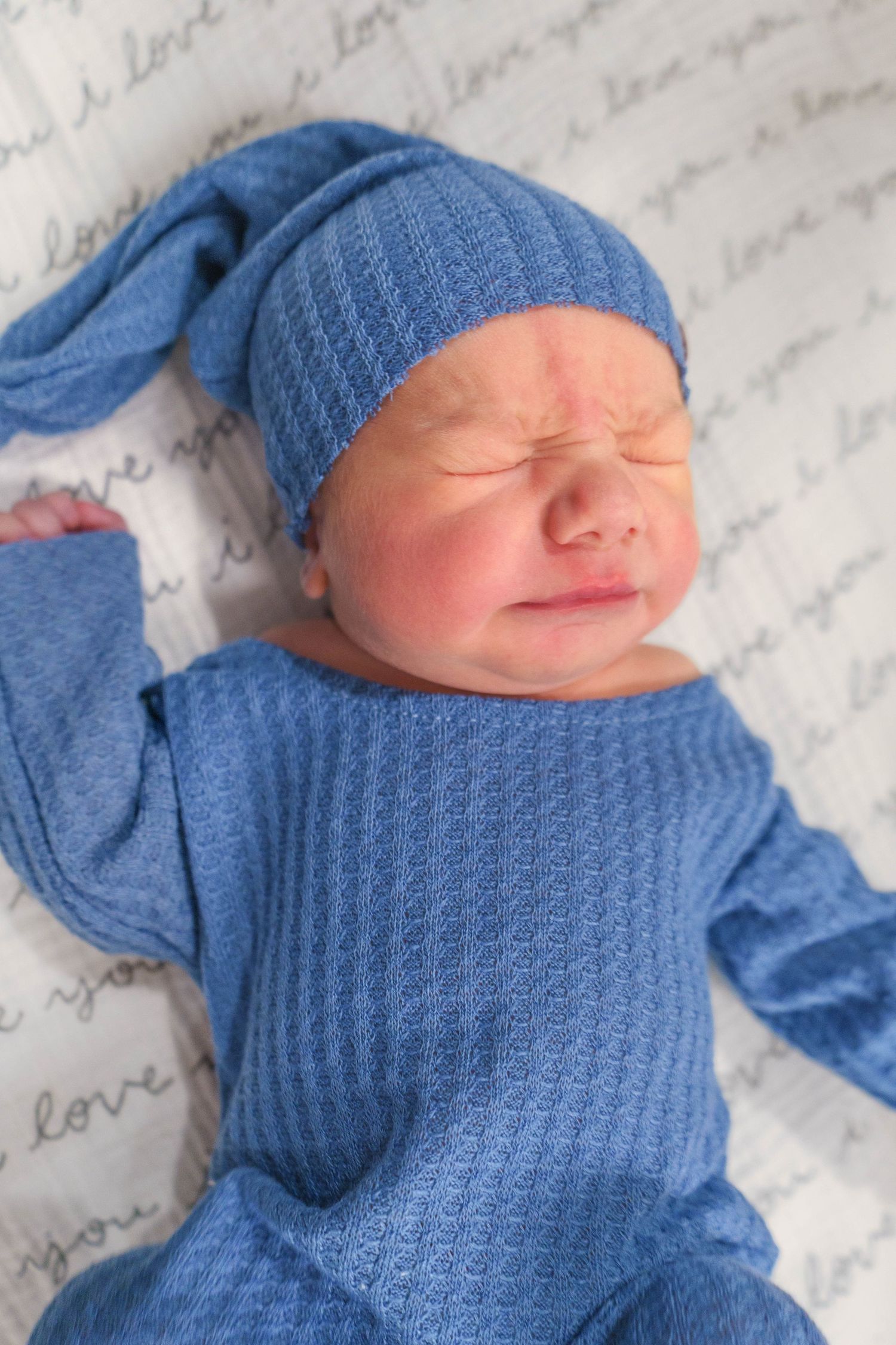 Newborn baby dressed in blue knit outfit and matching hat lying on love-printed blanket.