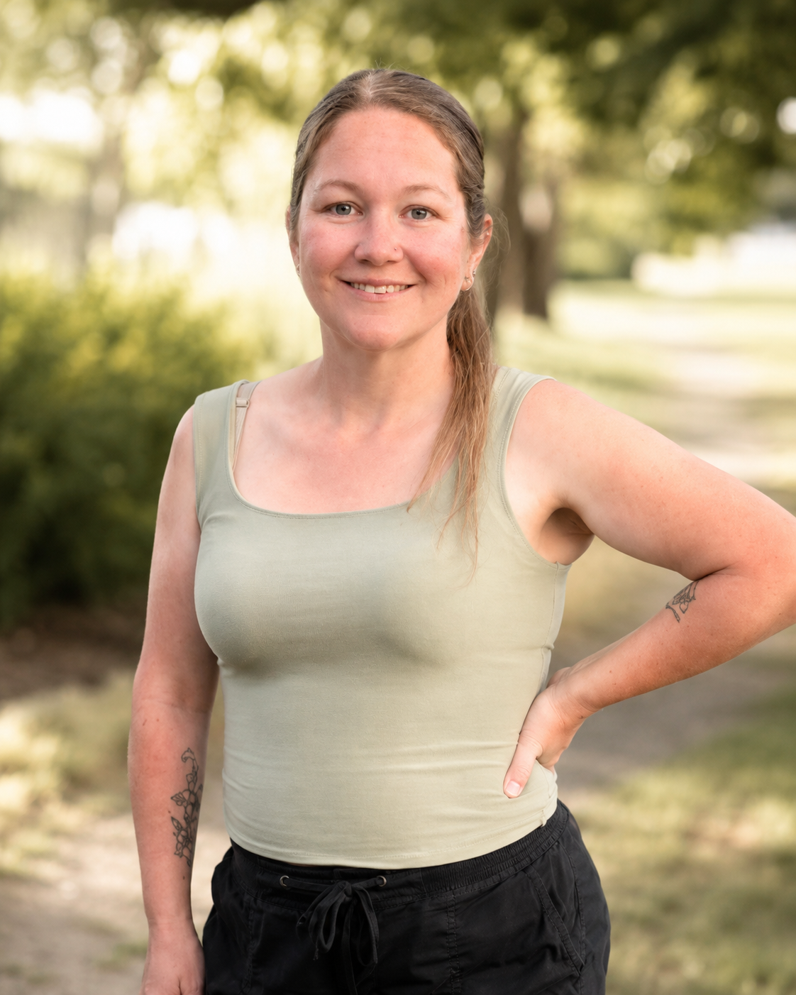 Smiling woman in sage green tank top posing outdoors with hand on hip, tattoos visible, trees in background.