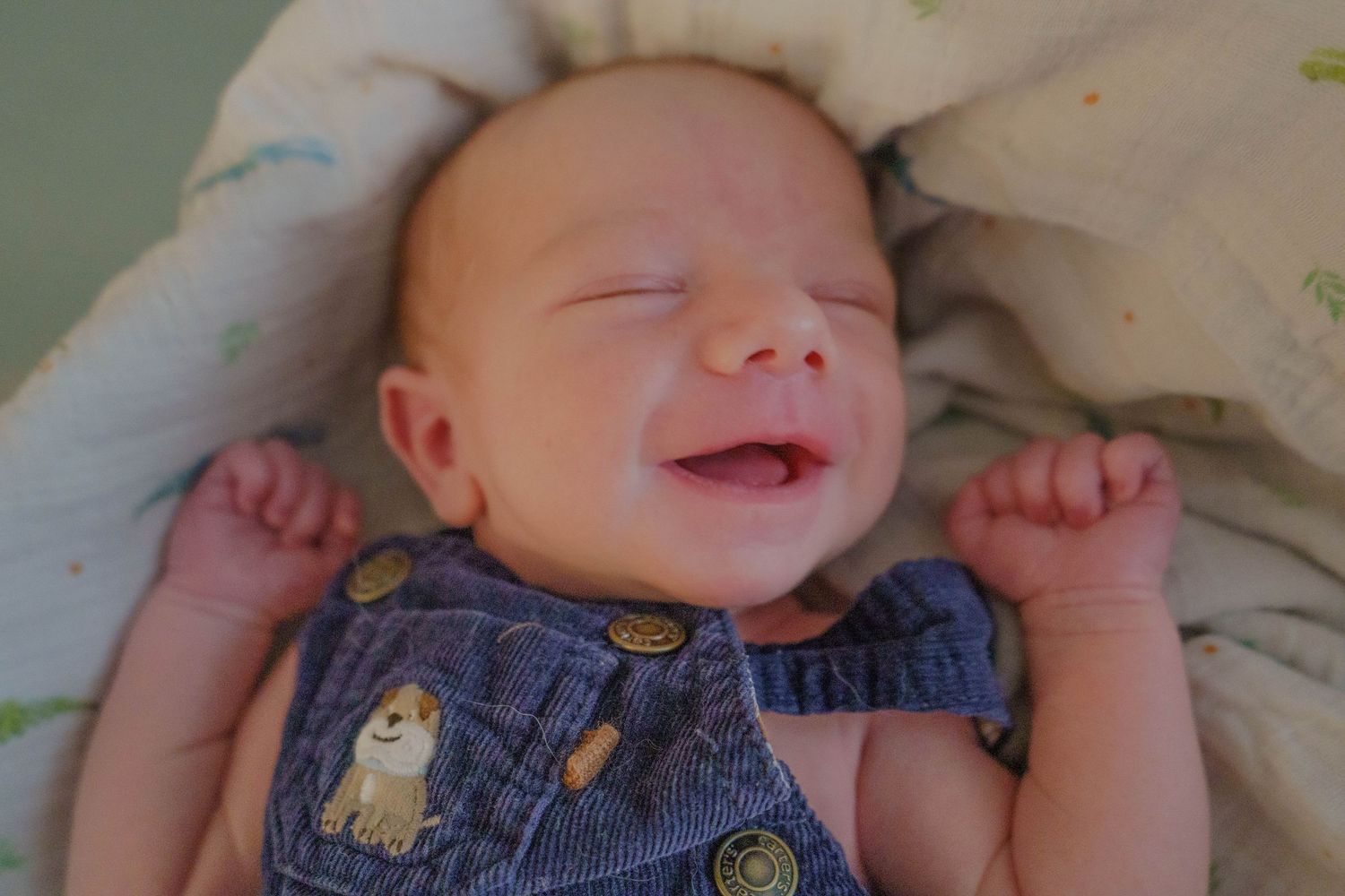 Smiling newborn baby in denim overalls lying on a soft blanket.