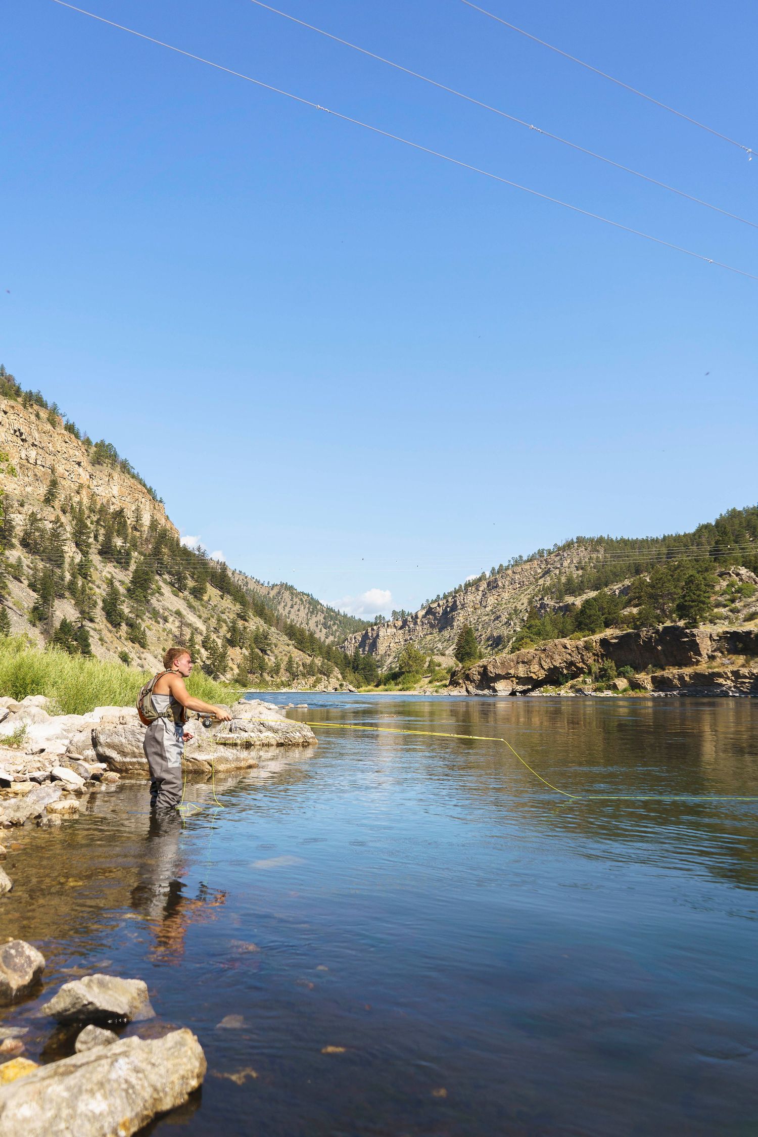 Fly fisherman wading in clear mountain river surrounded by rocky canyon landscape.
