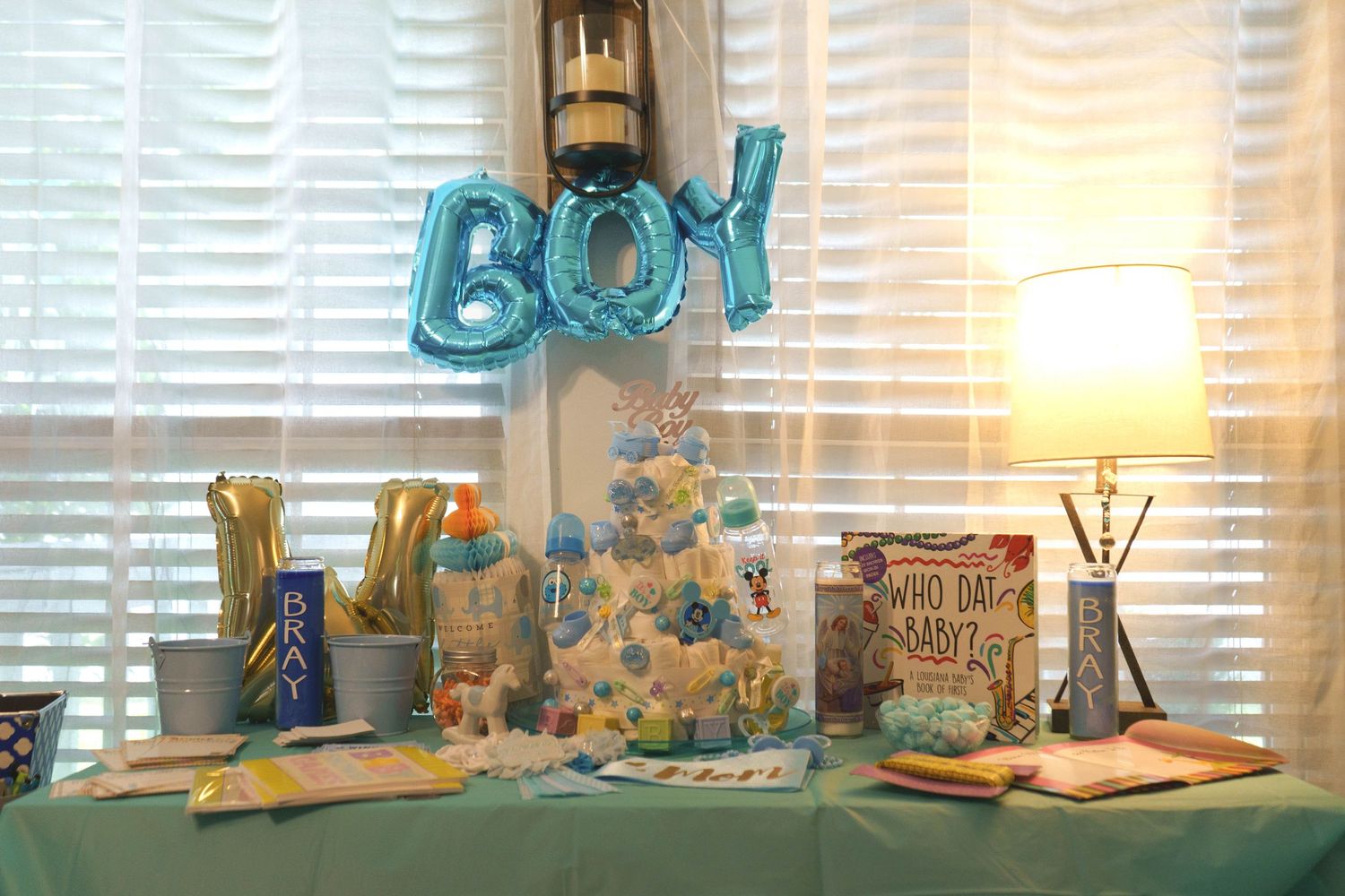 Baby boy shower table display with blue BOY balloons, decorated cake, gifts, and teal tablecloth decor.
