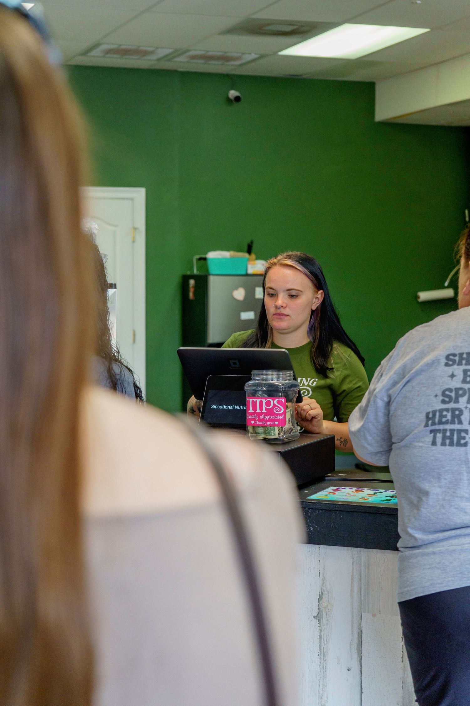 Female employee assisting customers at the counter of a nutrition smoothie bar with green walls.