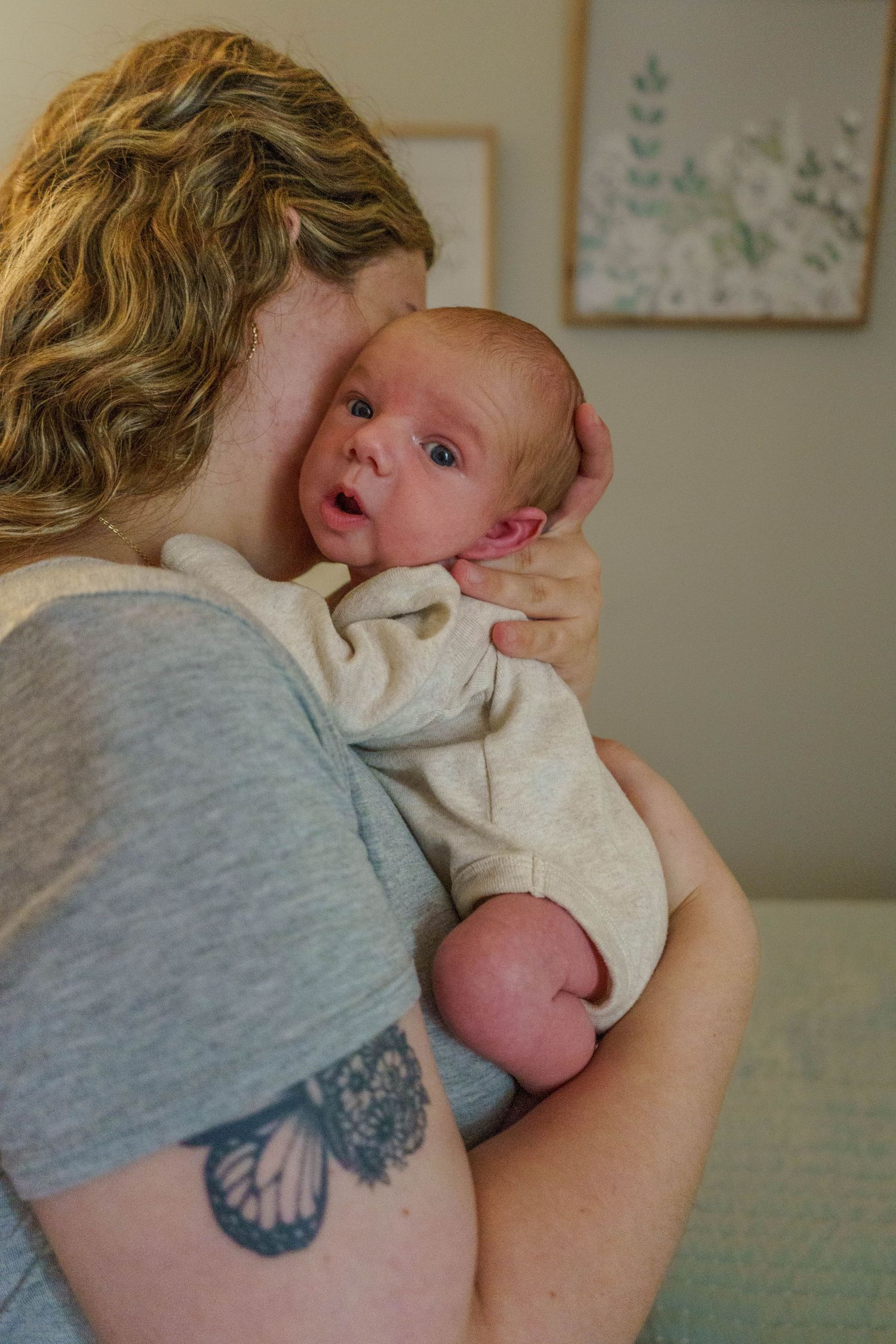 Mother with butterfly tattoo holding and kissing newborn baby in gray onesie.