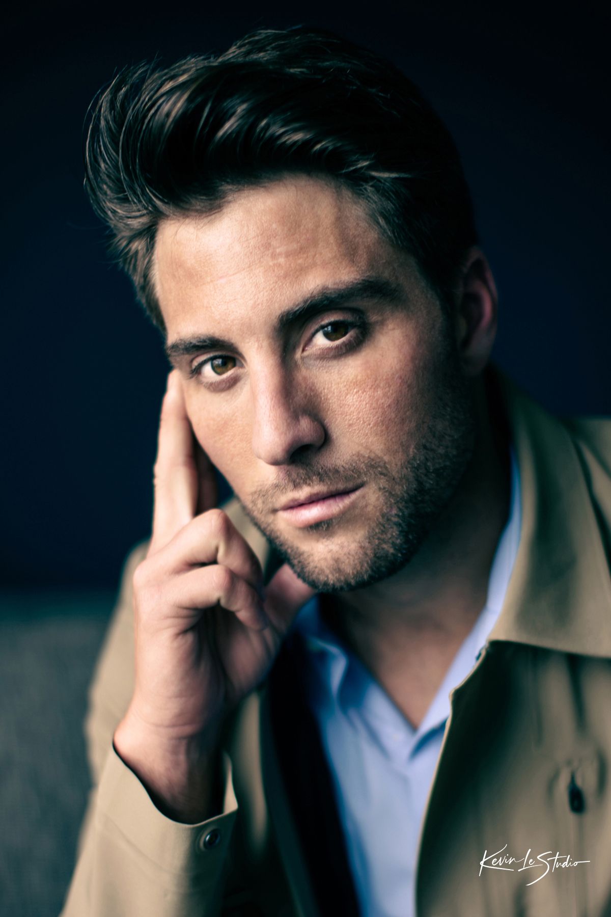 A young man with dark hair poses thoughtfully, resting his hand near his face, wearing a tan jacket and blue shirt.