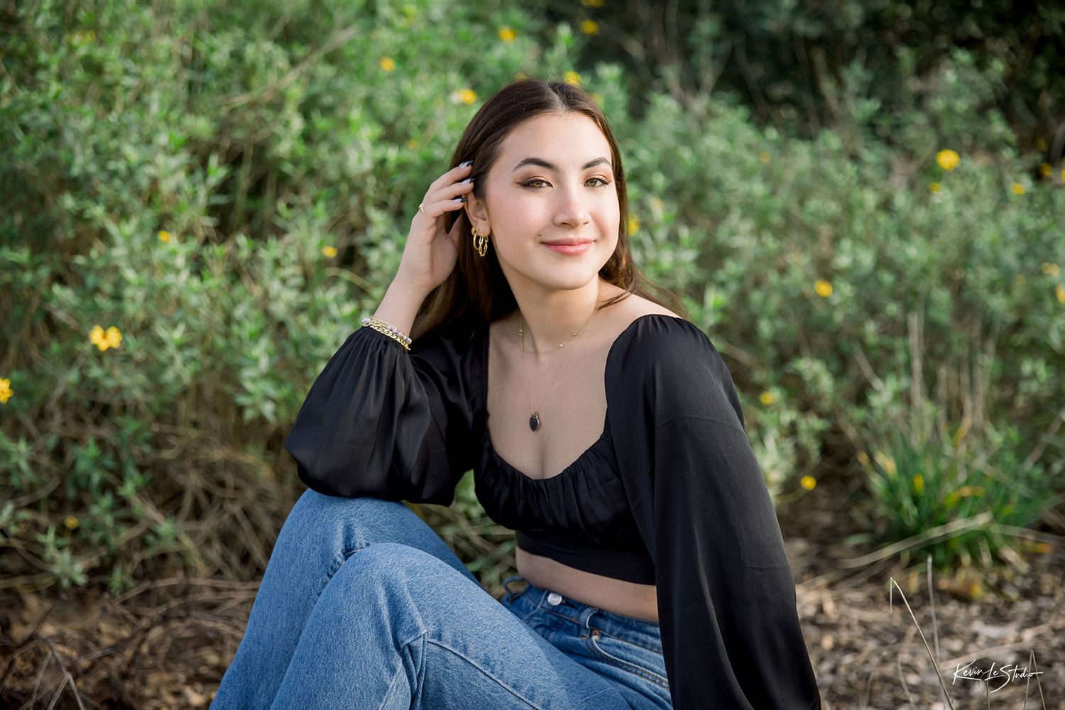 Young woman in black crop top and jeans sitting outdoors among green shrubs, posing for senior photos.