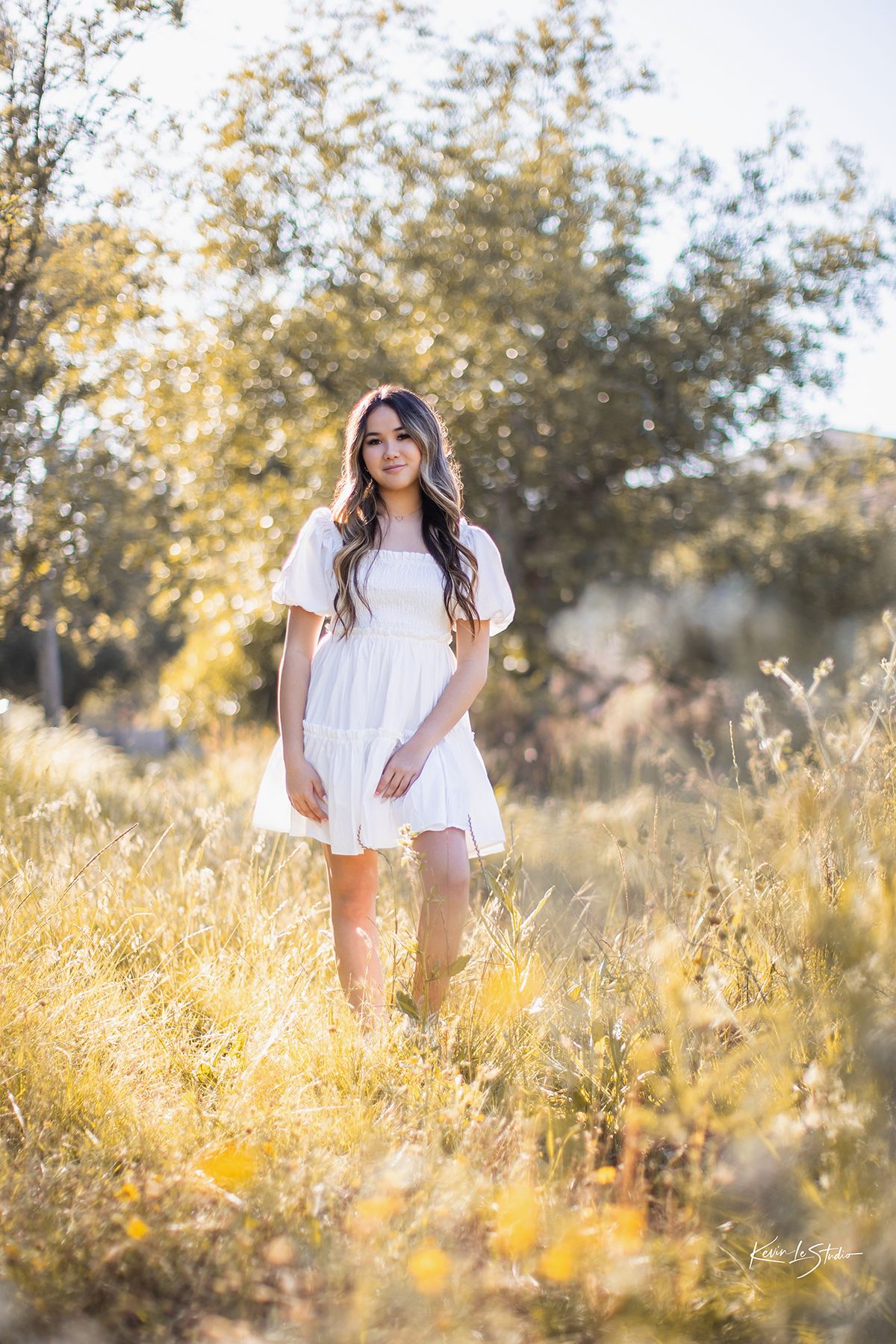 Brunette woman in white puff sleeve dress standing in glowing sunlit meadow with trees behind.