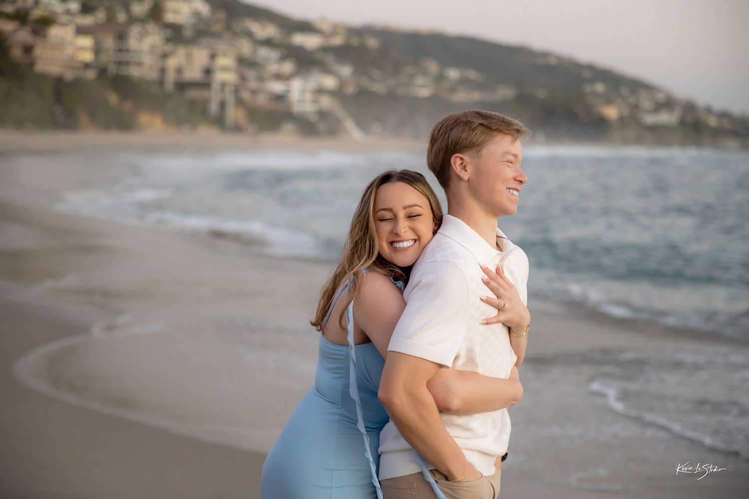 Joyful couple playing on sandy beach, woman hugging man from behind laughing.