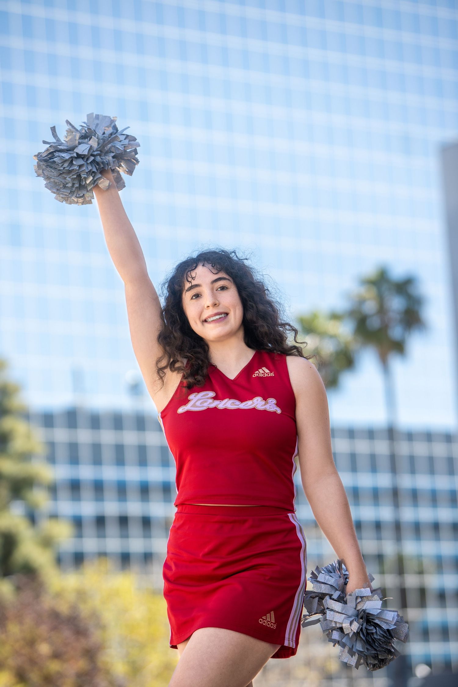 Cheerleader in red Lancers uniform raises silver pom-pom high in urban outdoor setting.